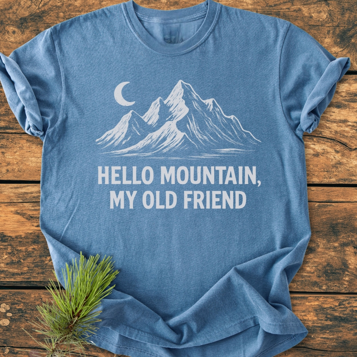 Hello Mountain My Old Friend - Vintage Wash Tee features a blue heather fabric with a white mountain and moon design, laid on wood, highlighting its relaxed fit and durable construction.