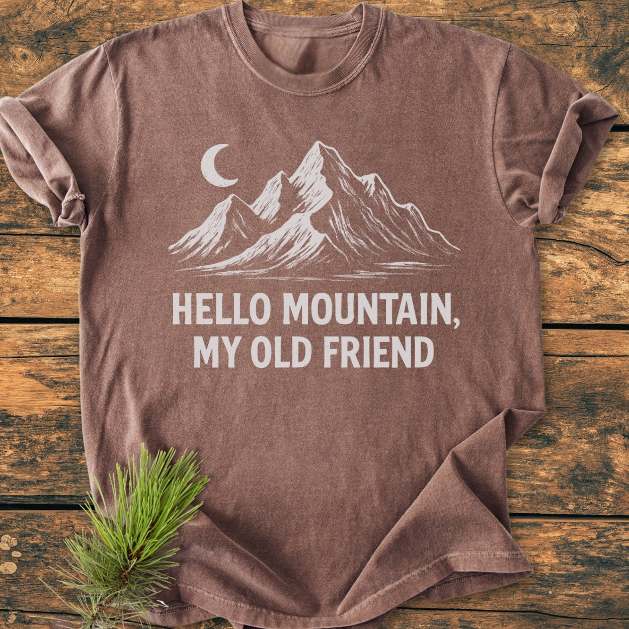 Hello Mountain My Old Friend - Vintage Wash Tee features a brown, short-sleeved design with a white mountain and crescent moon graphic, laid on a wooden surface with a pine sprig accent.