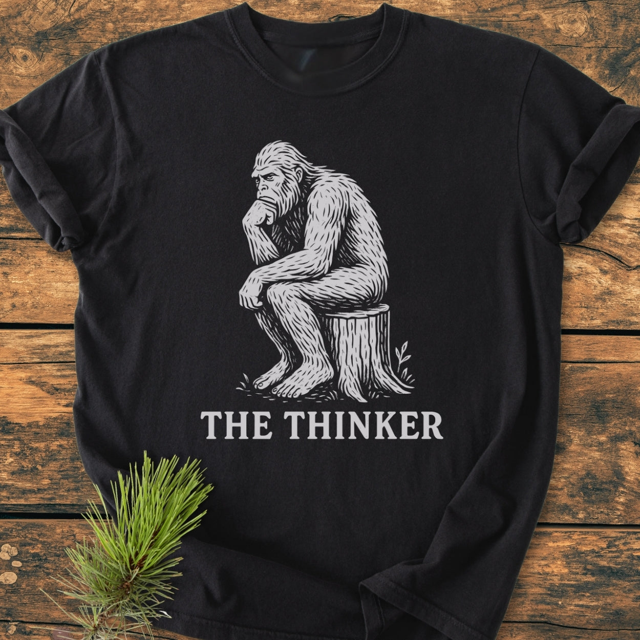 Bigfoot The Thinker - Vintage Wash Tee features a white illustration of a Bigfoot in thinker pose on a black background, laid on rustic wood, with THE THINKER caption below.