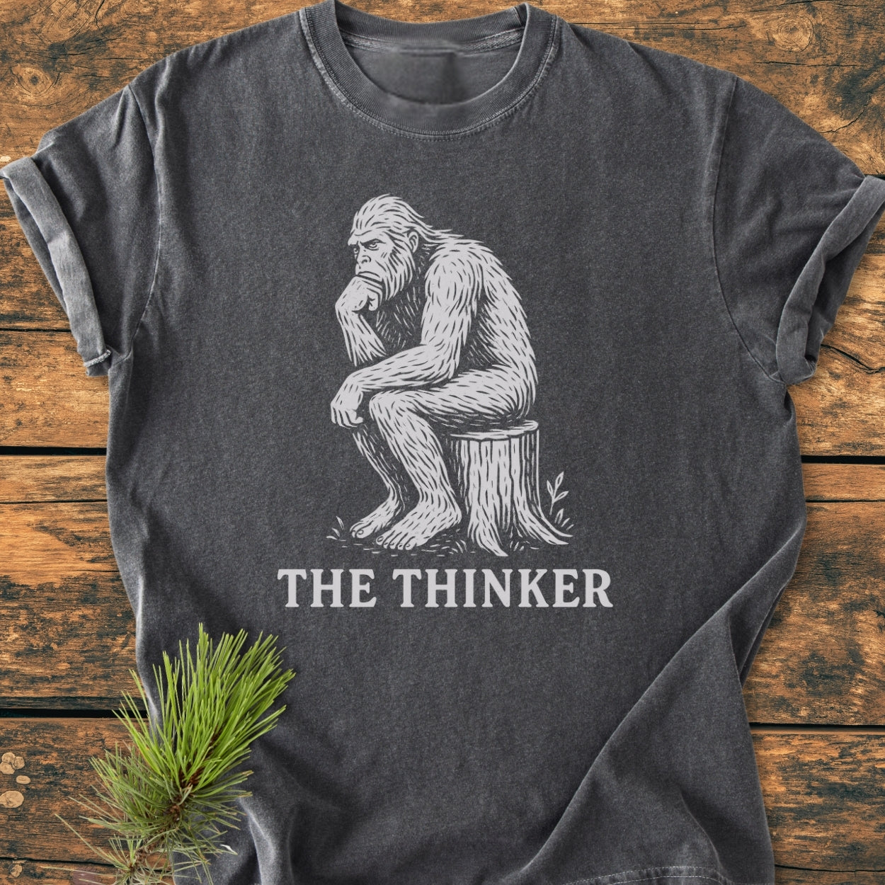 Bigfoot The Thinker - Vintage Wash Tee features a white Bigfoot illustration in a Thinker pose on a charcoal-grey shirt, laid on wood, with a pine sprig beside it.