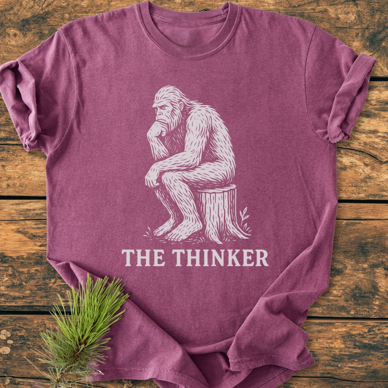 Bigfoot The Thinker - Vintage Wash Tee features a sketch of a Bigfoot in a thinker pose on a mauve shirt, laid on wood, with THE THINKER caption, offering comfort and durability.