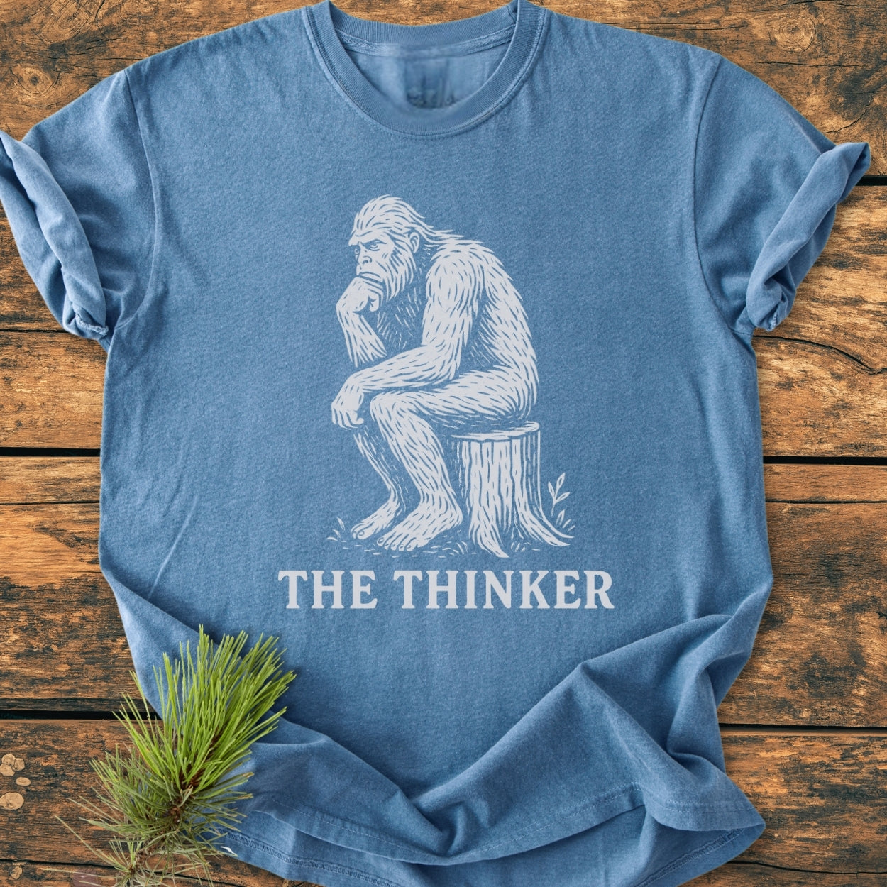 Bigfoot The Thinker - Vintage Wash Tee features a stylized ape in Thinker pose on a heather-blue T-shirt, laid flat with a pine sprig, highlighting its relaxed, durable design.