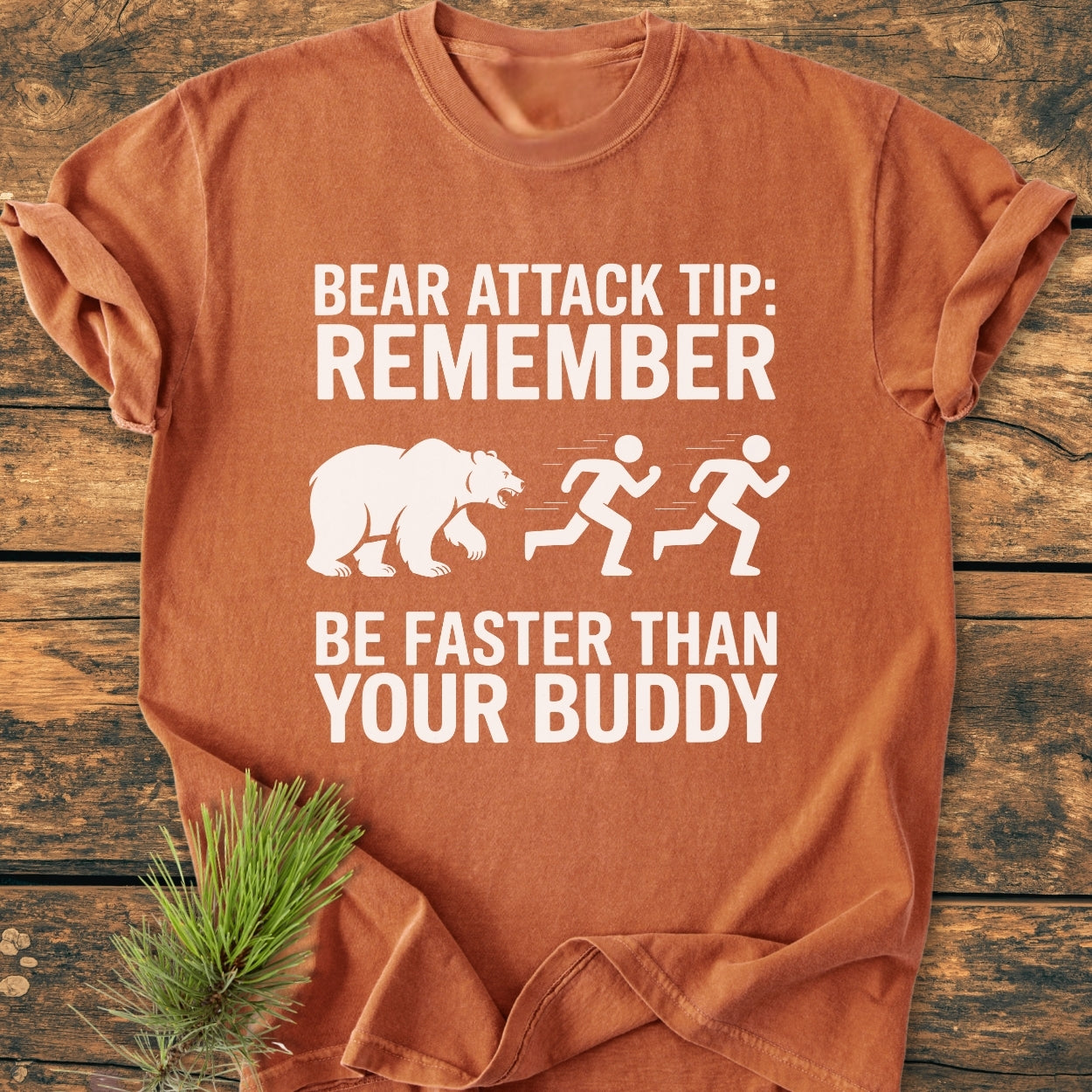 Bear Attack Tip Be Fast - Vintage Wash Tee, featuring a cartoon bear chasing stick figures with humorous text. Displayed on wooden surface with pine needles accentuating the garment’s rustic appeal.