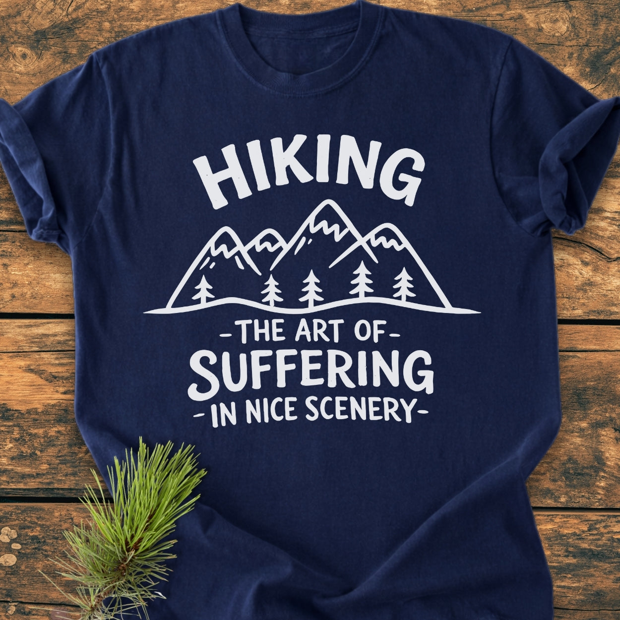 Navy-blue tee, “Hiking. The Art Of Suffering In Nice Scenery,” with mountain peaks and pine trees graphic, laid on wood. Pine twig accents the outdoorsy theme.