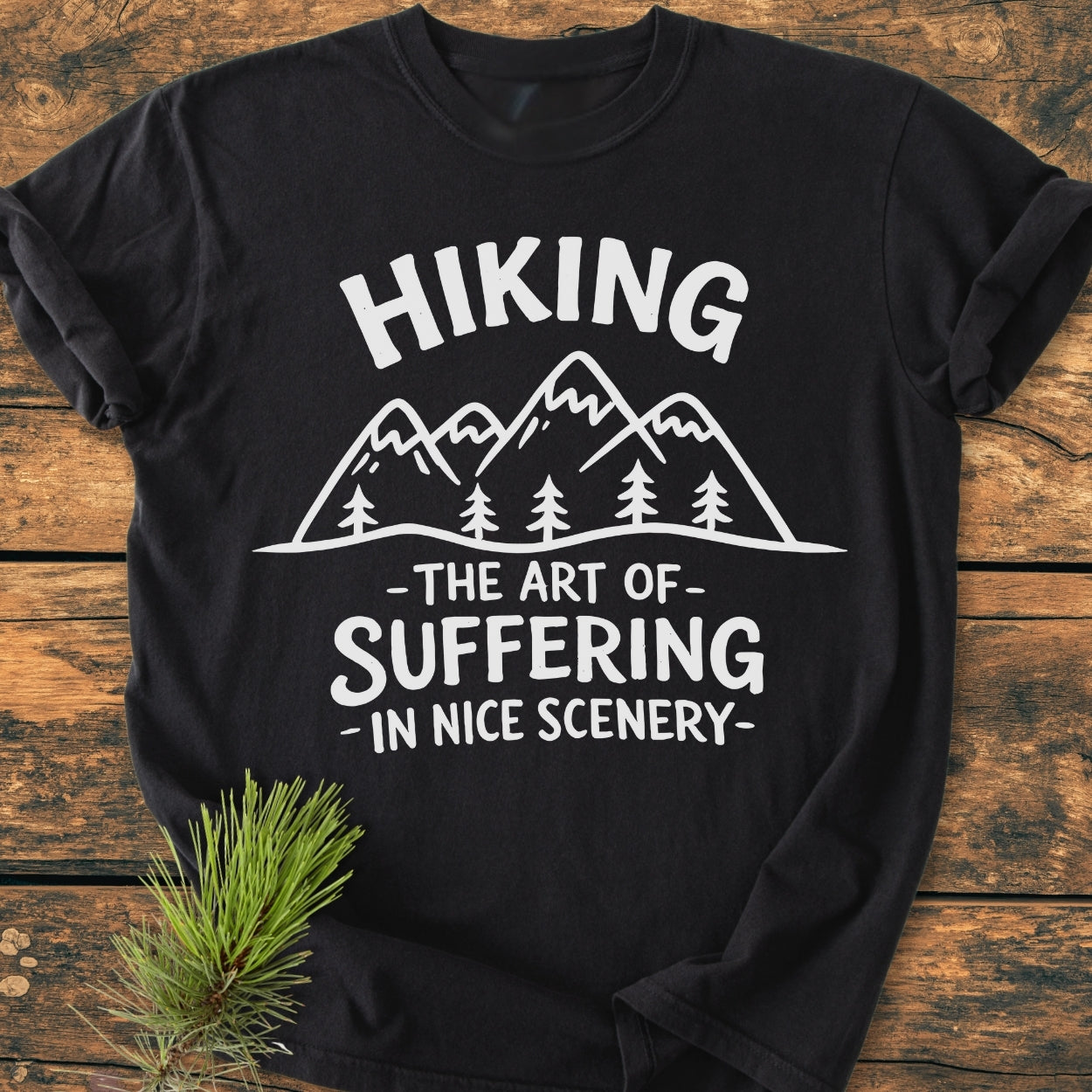 Black T-shirt on wood surface with Hiking. The Art Of Suffering In Nice Scenery graphic, featuring mountains and trees. Made from soft, durable, pre-shrunk 100% ring-spun cotton for comfort and a relaxed fit.