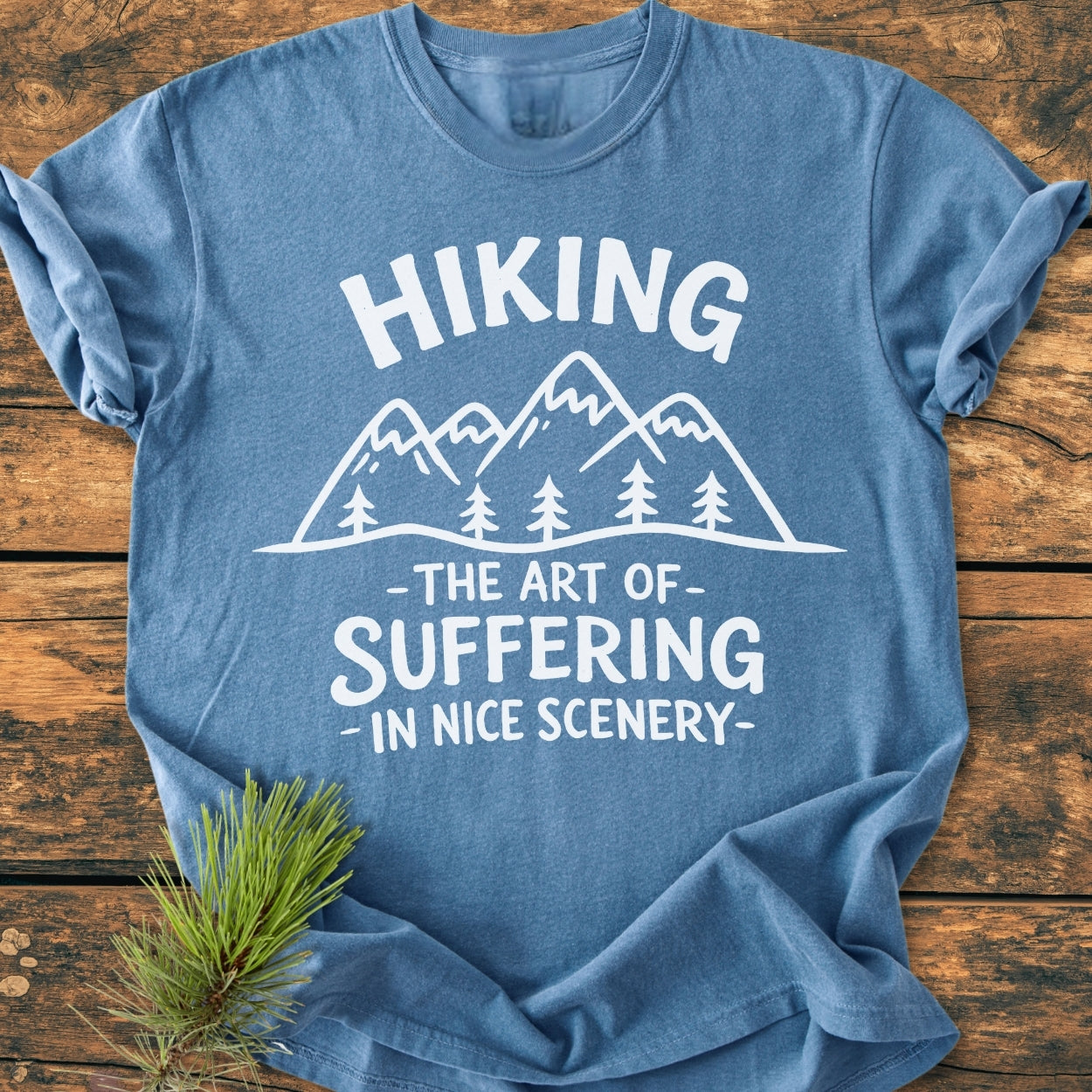 Heather-blue tee featuring a hand-drawn mountain graphic with pine trees and the slogan HIKING – The Art of Suffering in Nice Scenery, styled with a pine sprig beside it.