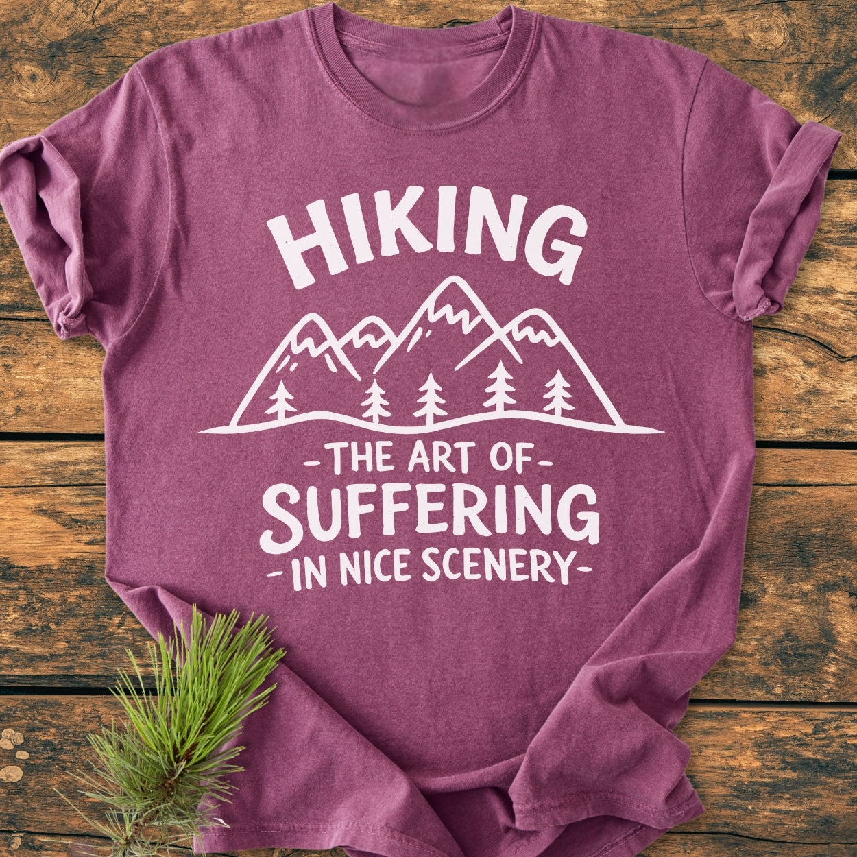 Heather-purple “Hiking. The Art Of Suffering In Nice Scenery - Vintage Wash Tee” featuring white mountain and pine line-art, laid folded on wood with pine needles beside.