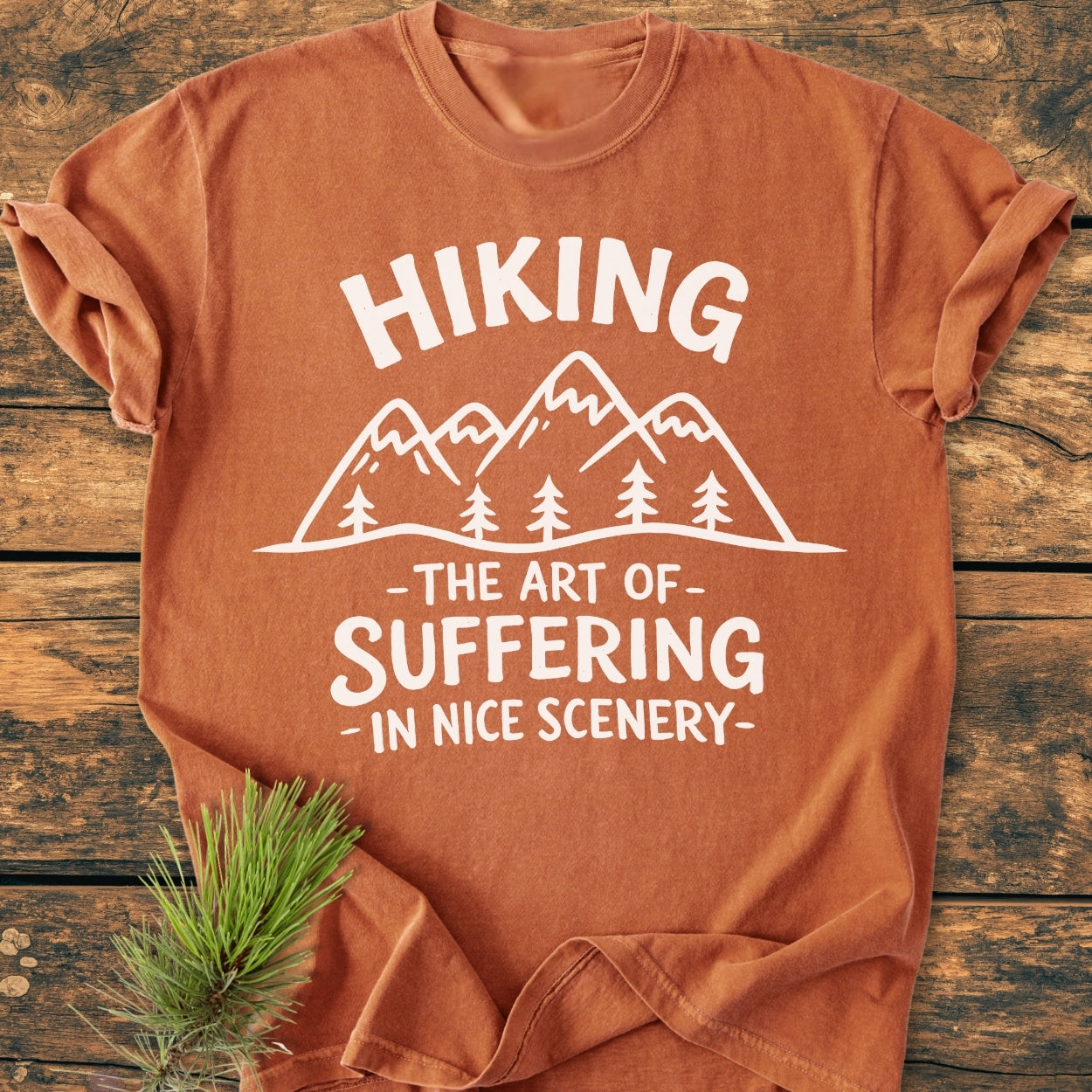 Hiking. The Art Of Suffering In Nice Scenery - Vintage Wash Tee featuring a mountain-and-pine-tree graphic on rust-orange fabric, styled with a pine sprig on a wooden surface for a rugged aesthetic.