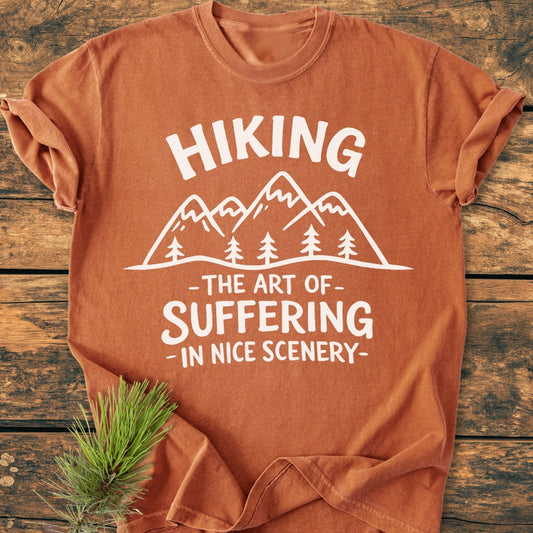 Hiking. The Art Of Suffering In Nice Scenery - Vintage Wash Tee featuring a mountain-and-pine-tree graphic on rust-orange fabric, styled with a pine sprig on a wooden surface for a rugged aesthetic.