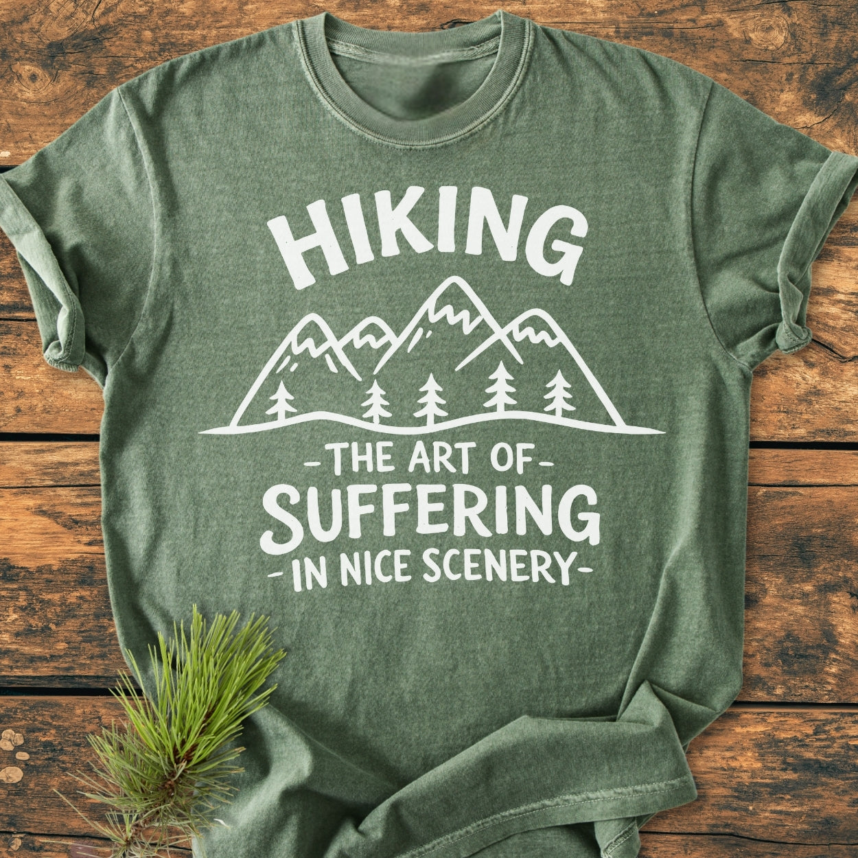 Heather-green T-shirt with white mountain and pine drawing, slogan Hiking—The Art Of Suffering—In Nice Scenery. Laid flat with fir sprig, emphasizing comfort and relaxed fit.