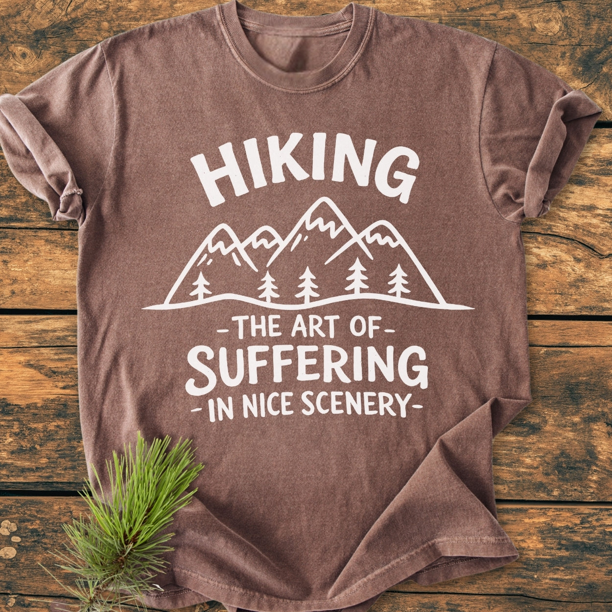 Hiking. The Art Of Suffering In Nice Scenery - Vintage Wash Tee features a mountain graphic on a brown short-sleeve shirt, laid on wood, with evergreen needles accentuating the outdoor theme.