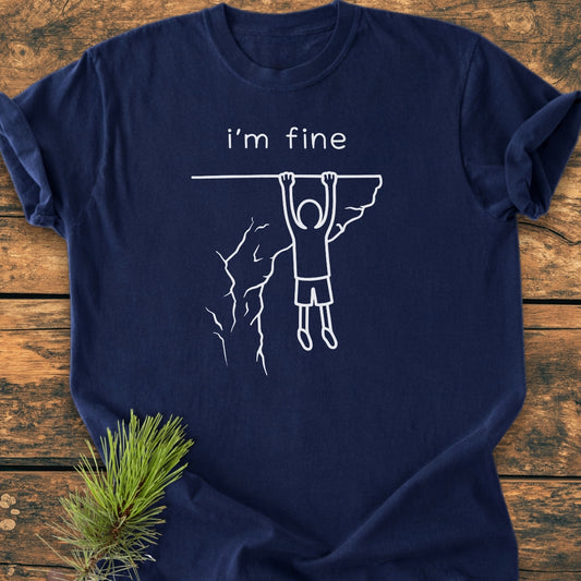 I'm Fine - Vintage Wash Tee featuring a stick-figure cliffhanger design with i’m fine. text, laid on a wooden surface with a pine sprig. Made of soft-washed, durable 100% ring-spun cotton.