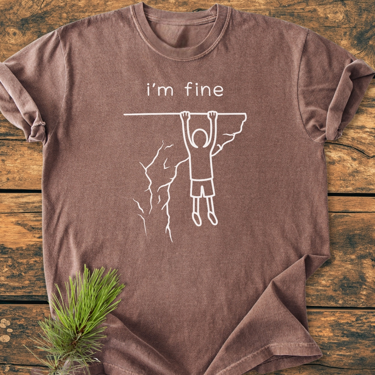 I'm Fine - Vintage Wash Tee features a stick-figure cliffhanger design and 'i'm fine' text on a soft, garment-dyed brown T-shirt, laid on wood with pine needles accent.