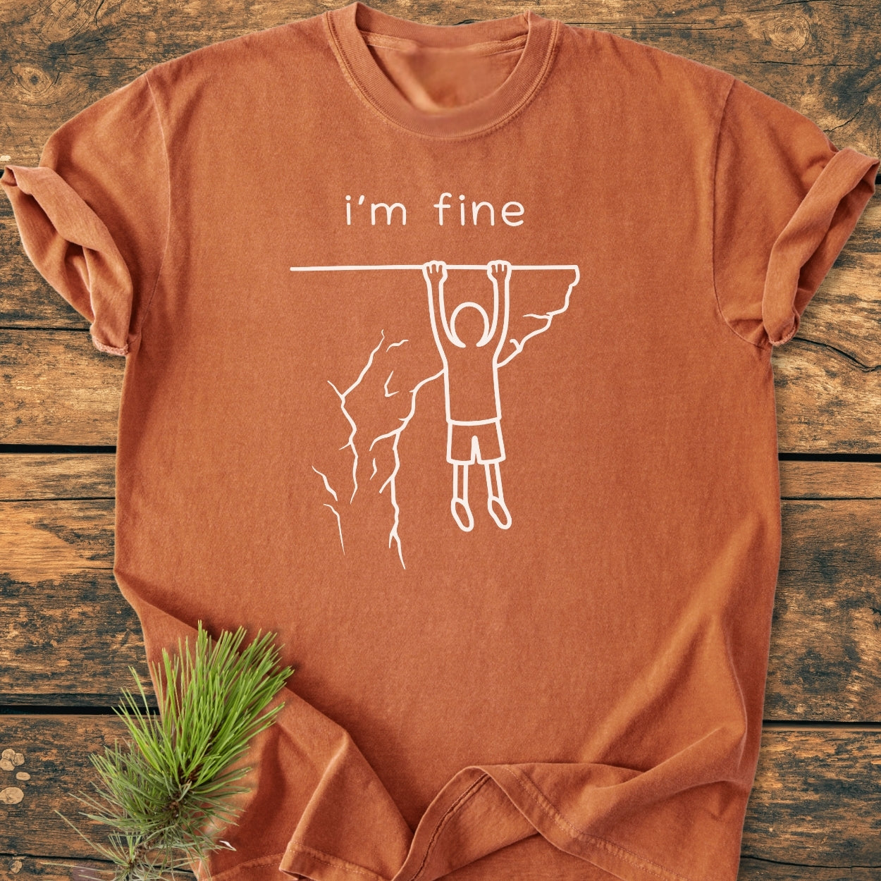 I'm Fine - Vintage Wash Tee in burnt sienna features a white print of i’m fine and a stick-figure clinging to a cliff. Laid flat on wood, with an evergreen sprig beside.
