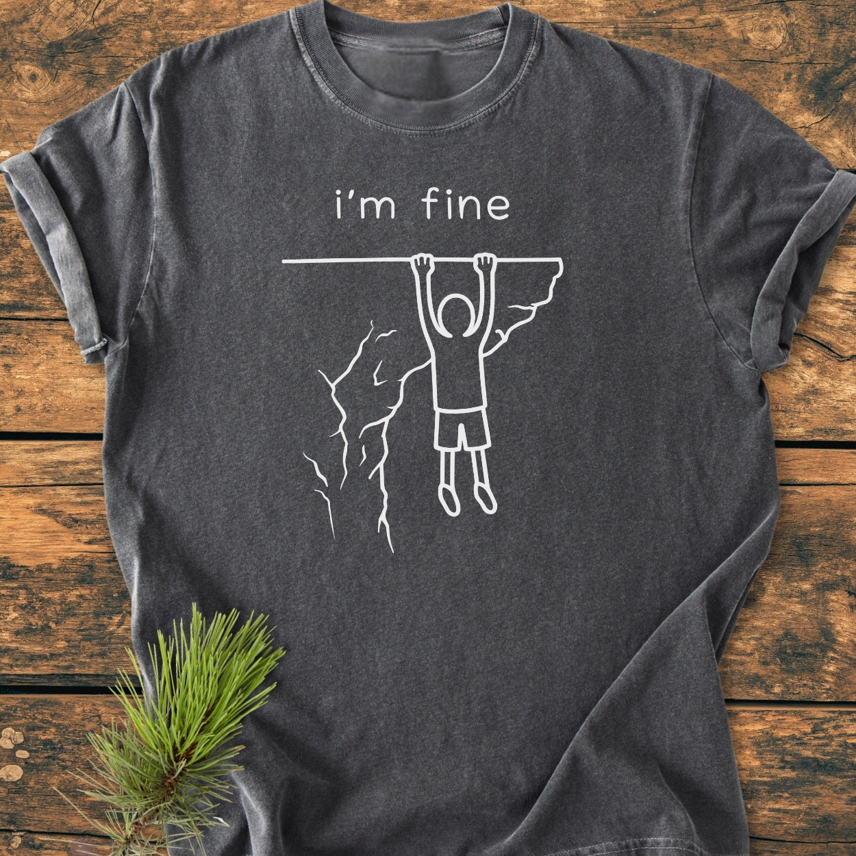 The I'm Fine - Vintage Wash Tee features a white stick-figure design and text on a charcoal-gray background, laid on rustic wood with a pine sprig, highlighting its unique and relaxed style.
