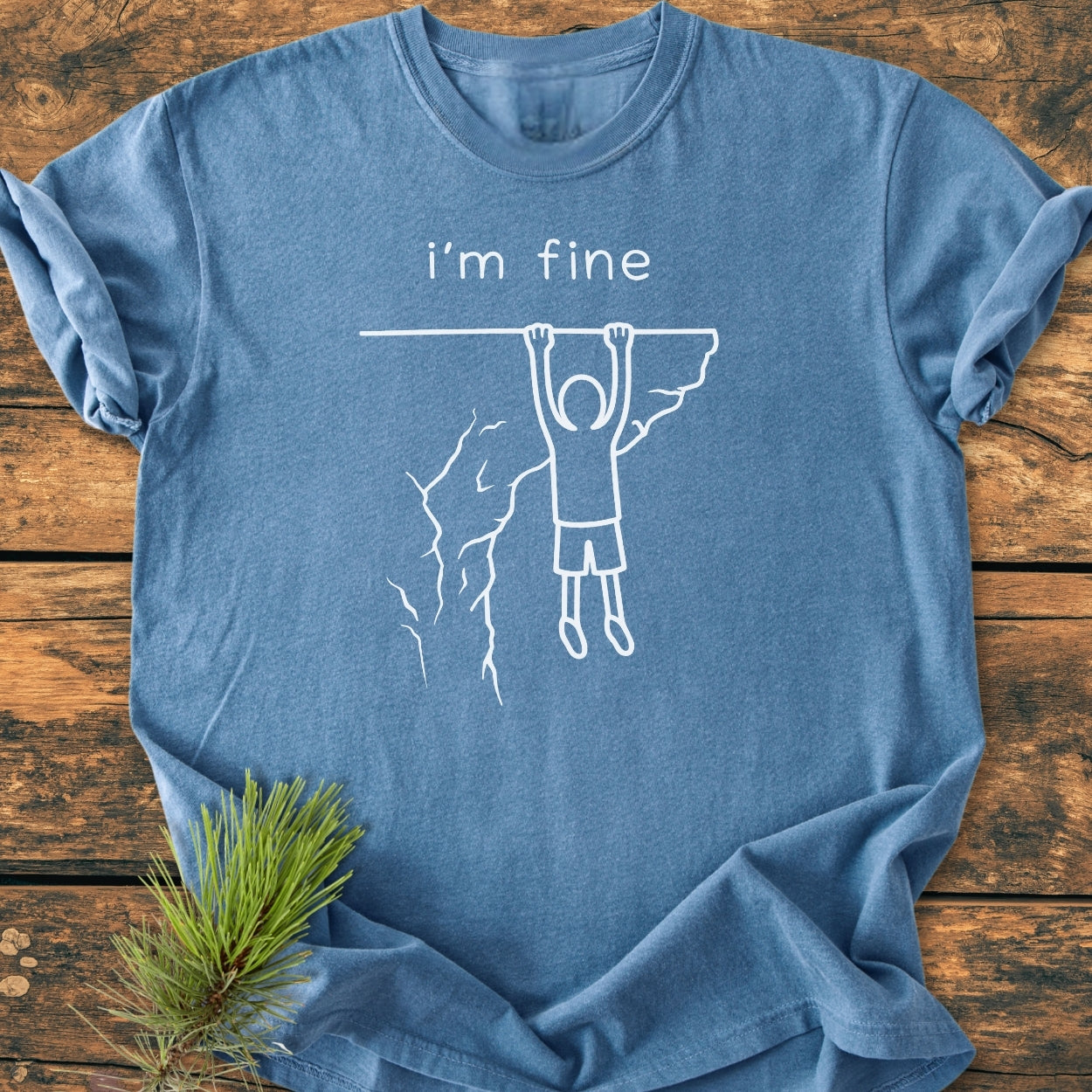 I'm Fine - Vintage Wash Tee featuring a stick-figure cliffhanger graphic with “i’m fine.” text, laid flat on a wooden surface, with a small pine sprig beside it.