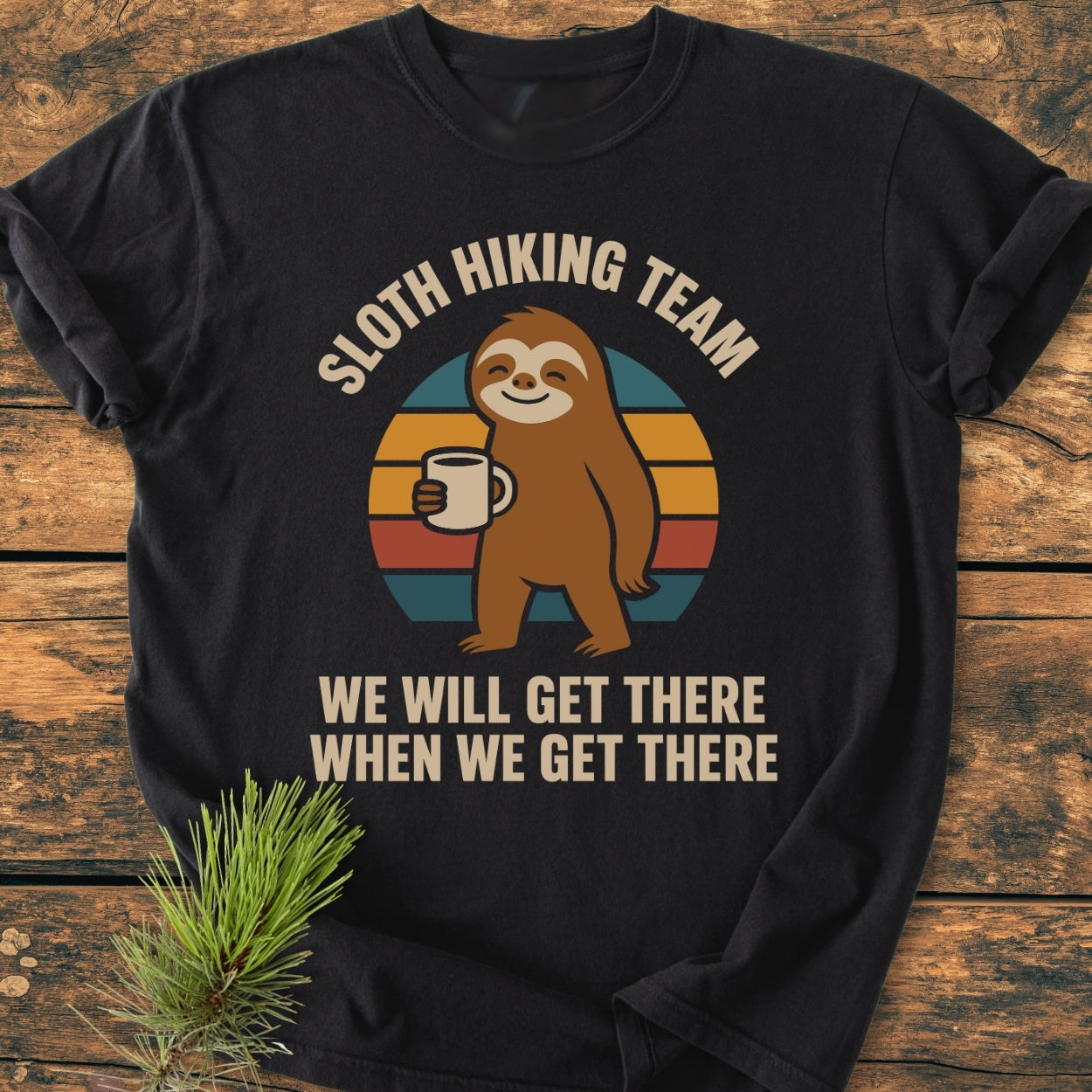 Sloth Hiking Team - Vintage Wash Tee featuring a cartoon sloth with a coffee mug on a retro-striped circle, laid on a wooden surface. Text reads, “WE WILL GET THERE WHEN WE GET THERE.”