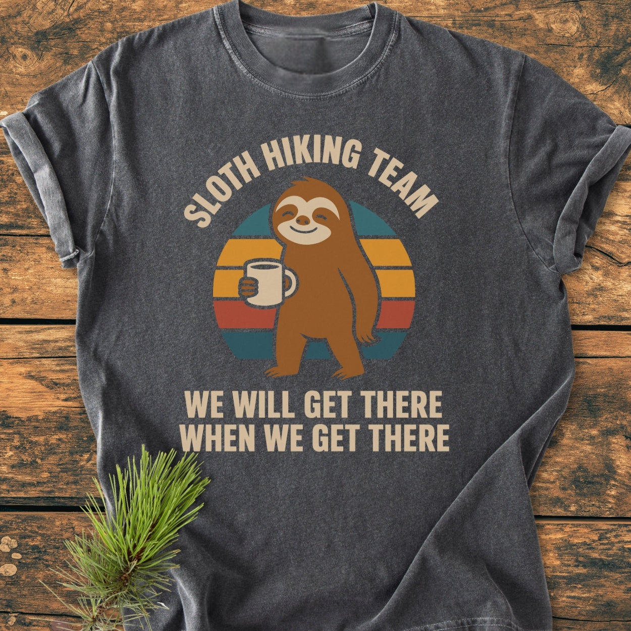 Sloth Hiking Team - Vintage Wash Tee featuring a cartoon sloth with a coffee mug on a charcoal-gray, soft-washed fabric, styled with a pine sprig on a wooden surface.