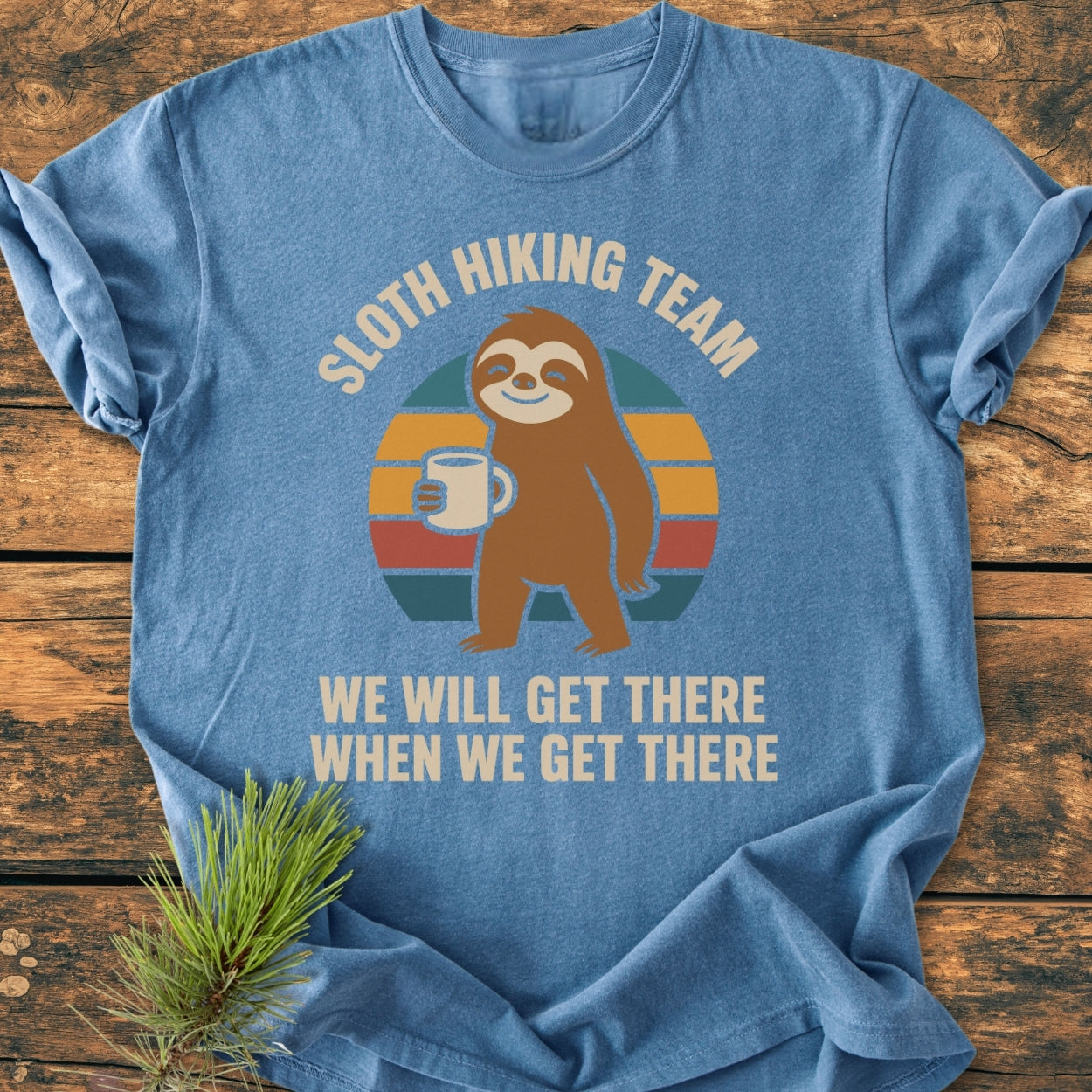 Sloth Hiking Team - Vintage Wash Tee features a cartoon sloth with a mug against a stylized sunset, laid on wood with a pine branch. Cozy, durable, relaxed fit in heather-blue.