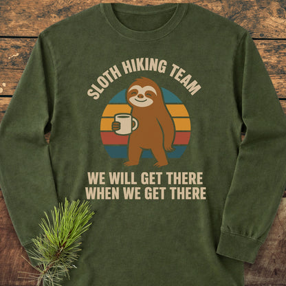 Sloth Hiking Team Long-Sleeve Tee featuring a cartoon sloth with a coffee mug on a retro-stripe circle, surrounded by playful text, laid on wood with pine accents. Crafted from soft, garment-dyed cotton.
