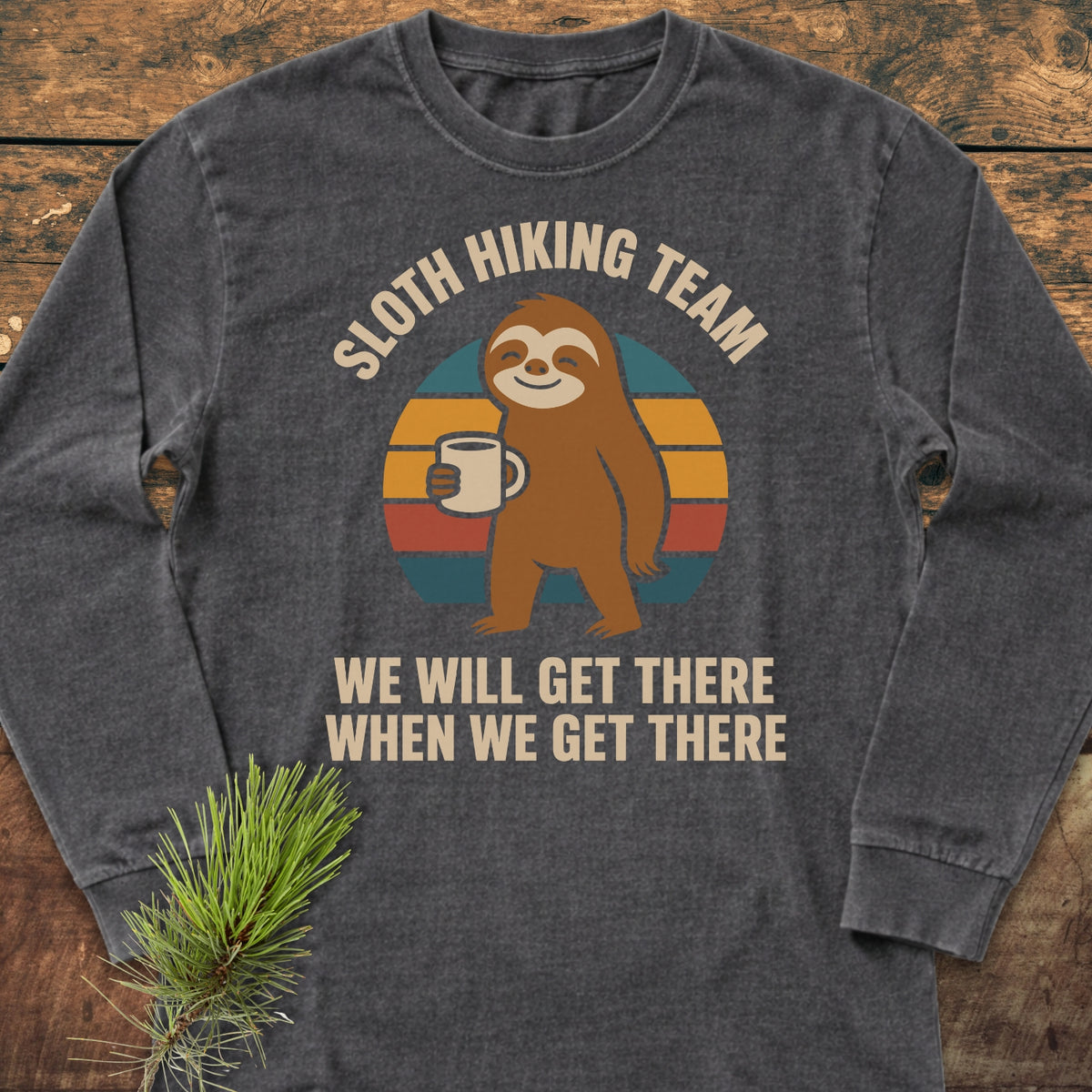Sloth Hiking Team Long-Sleeve Tee featuring a cartoon sloth with a mug on a striped circle, WE WILL GET THERE WHEN WE GET THERE text, on dark-gray fabric, laid on wood.