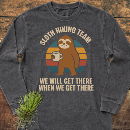Sloth Hiking Team Long-Sleeve Tee featuring a cartoon sloth with a mug on a striped circle, WE WILL GET THERE WHEN WE GET THERE text, on dark-gray fabric, laid on wood.