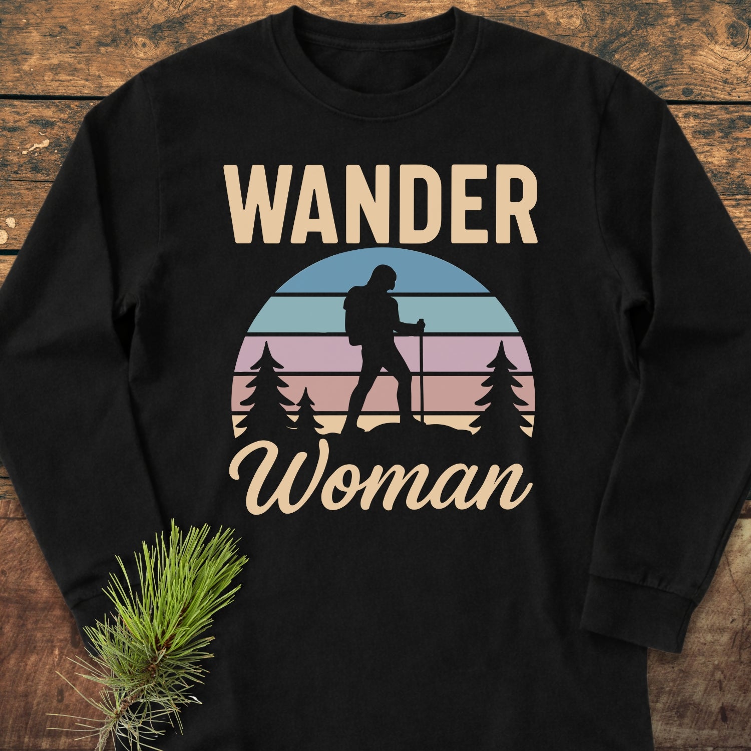 Wander Woman Retro Long-Sleeve Tee features a stylized hiker under a sunrise graphic with WANDER above and Woman below. Laid on wood with a pine sprig, it's cozy 100% cotton.