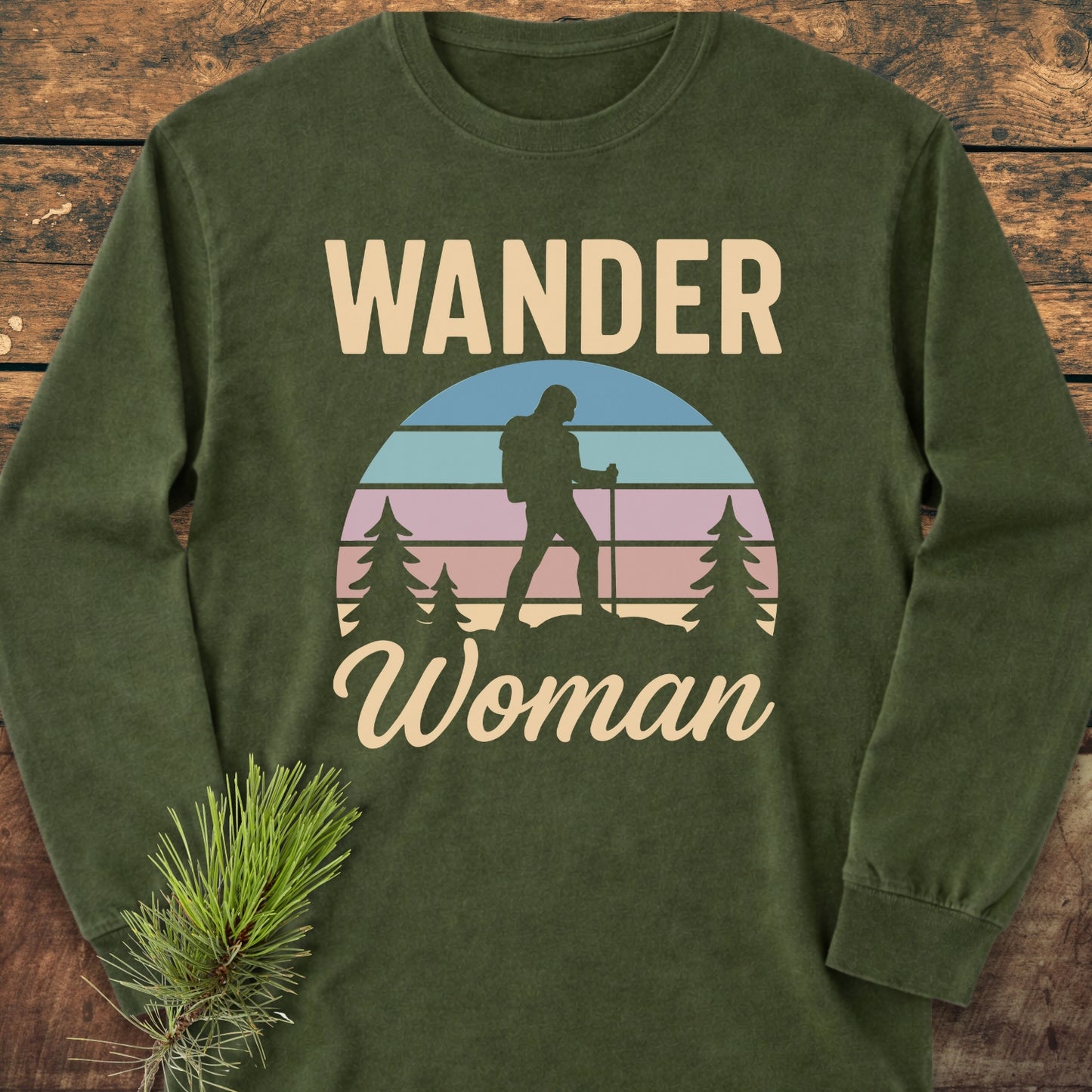 Wander Woman Retro Long-Sleeve Tee featuring a circular graphic of a woman hiker with trekking poles, surrounded by pine trees, printed on an olive-green, garment-dyed cotton tee for a relaxed fit.