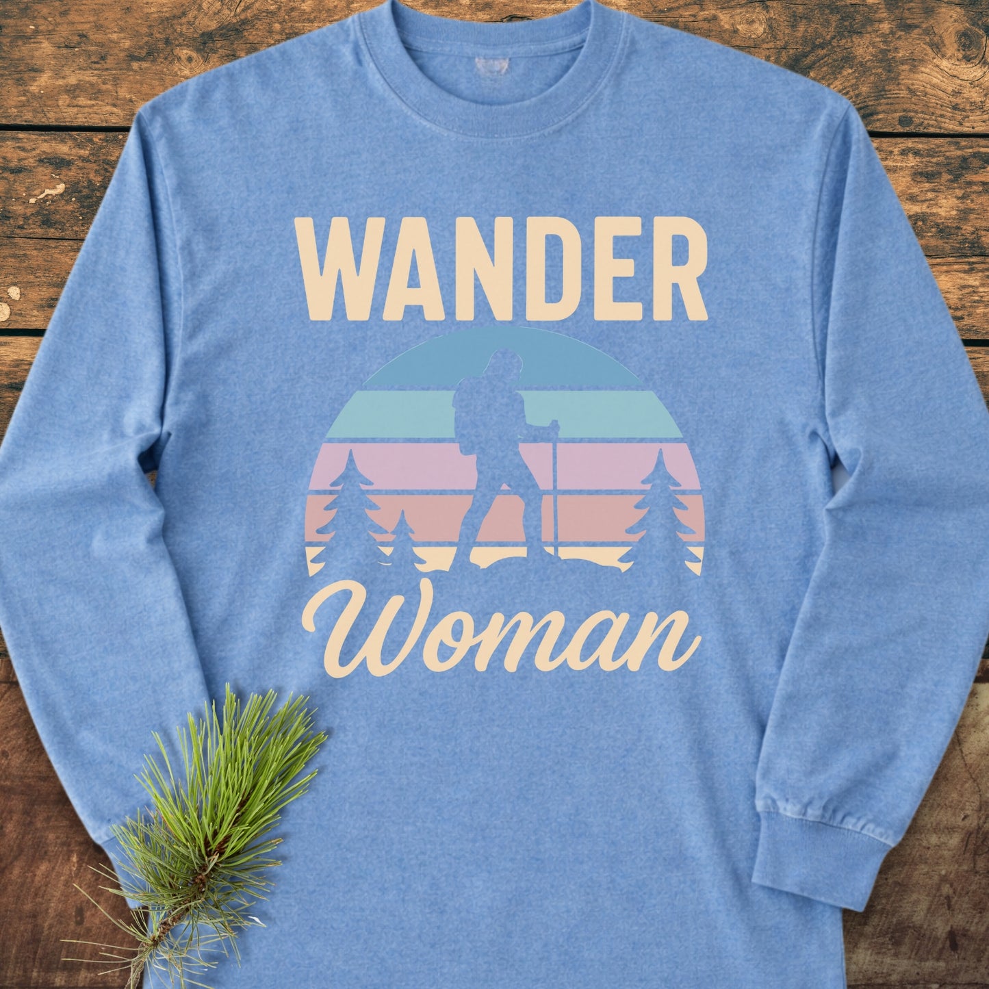 Wander Woman Retro Long-Sleeve Tee featuring a silhouetted hiker and pine trees graphic, crafted from soft 100% ring-spun cotton for a relaxed fit, displayed on a wooden surface with a pine branch.