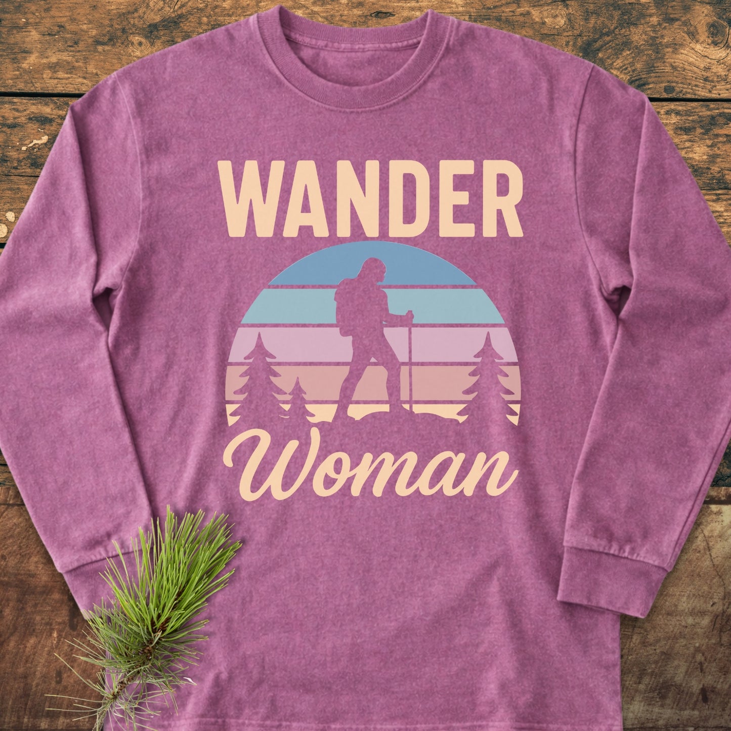 Wander Woman Retro Long-Sleeve Tee features a mauve-purple hue with a cream and pastel graphic of a hiker silhouette against a retro sunset motif, laid on rustic wood.