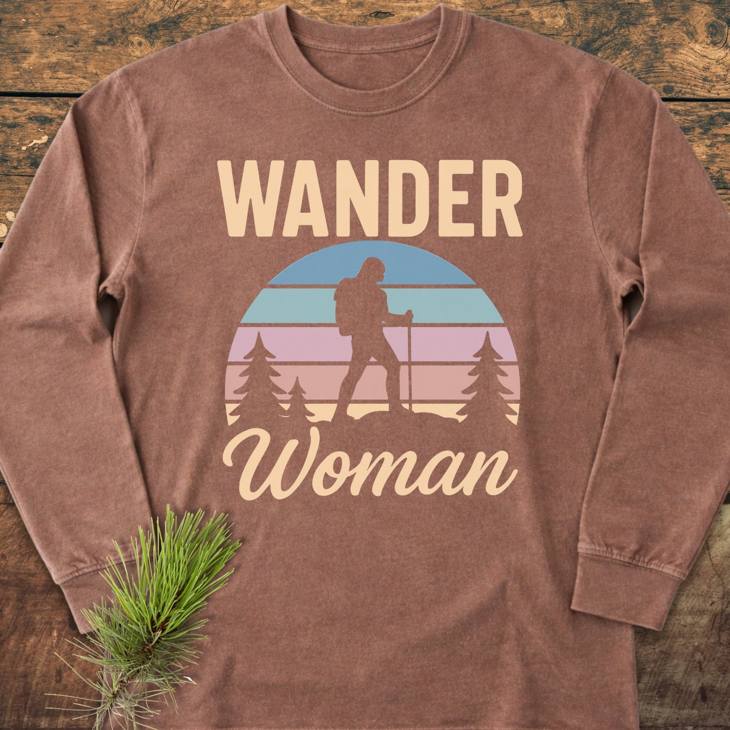 Wander Woman Retro Long-Sleeve Tee featuring a hiker silhouette against a stylized sunset, surrounded by pine trees. Laid on wood with a pine sprig prop, highlighting its soft, 100% ring-spun cotton fabric.