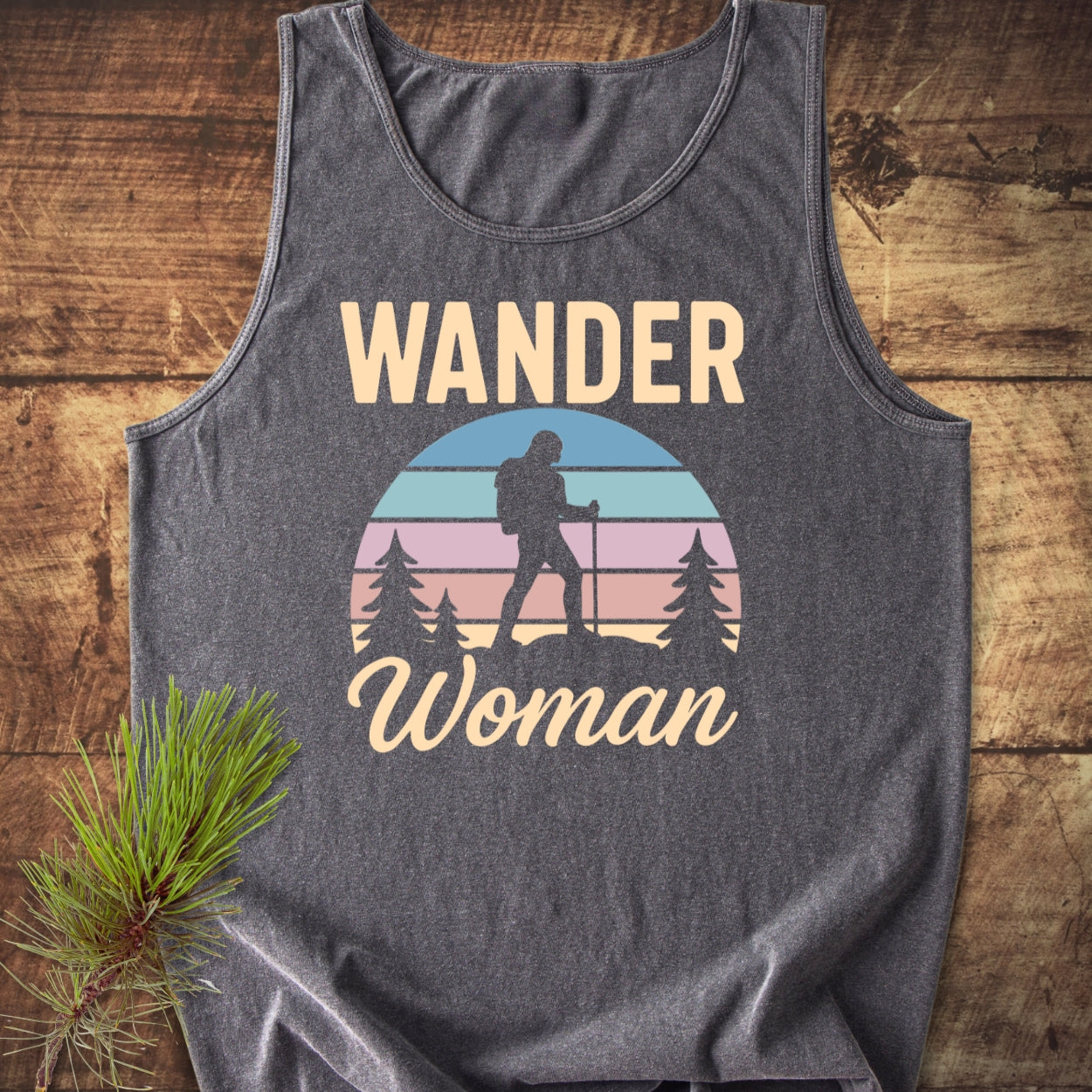 Wander Woman Retro Tank Top featuring a hiking silhouette against a pastel striped sunset, with pine trees and slogan, on a heather-gray sleeveless design, laid on wood beside a pine sprig.