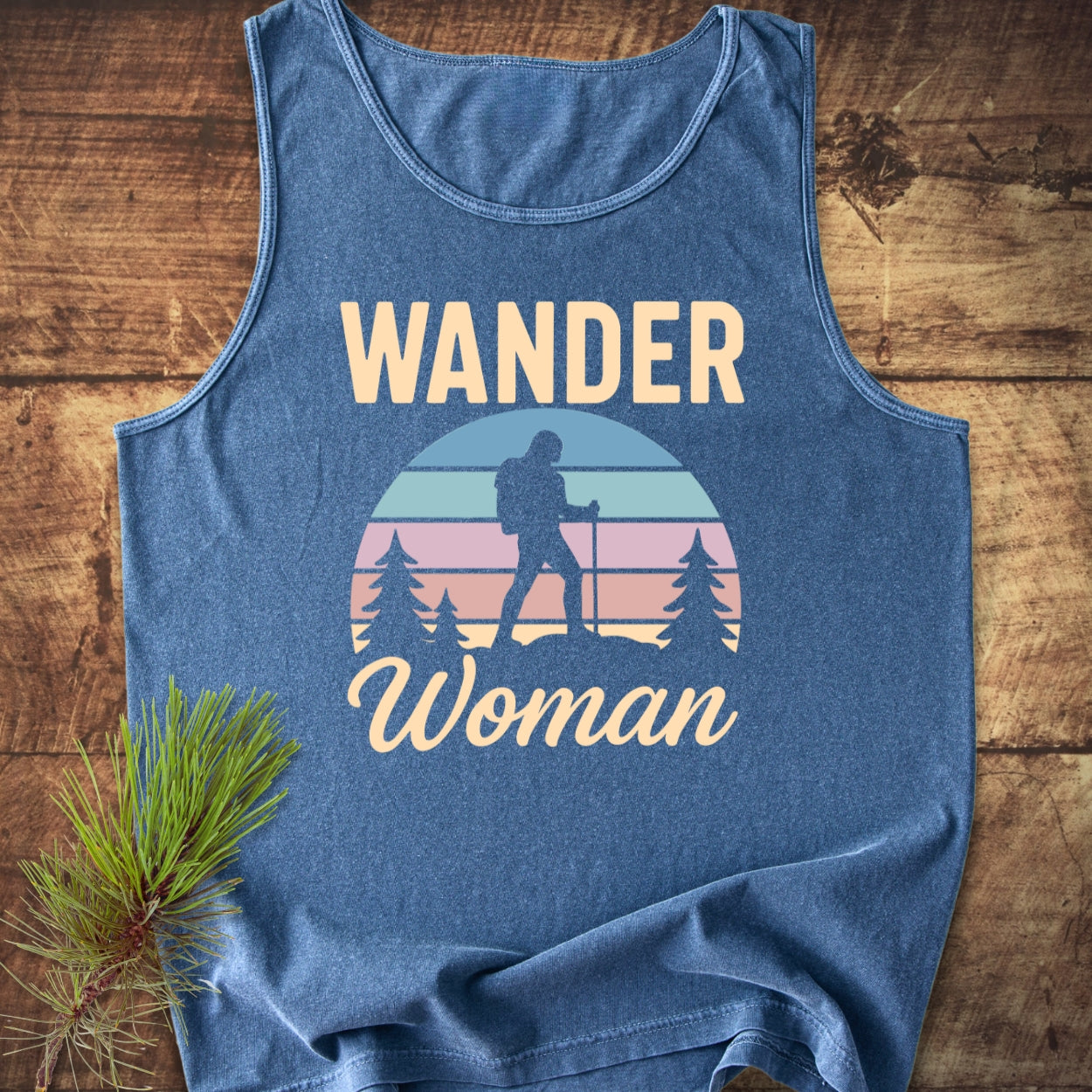 Wander Woman Retro Tank Top featuring a female hiker graphic against a sunset backdrop with pine trees, conveying an outdoor theme. Laid on a wooden surface, complemented by a pine branch.