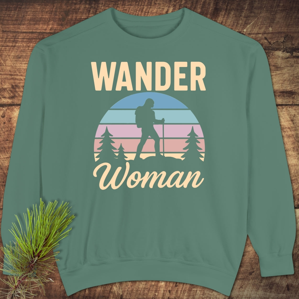 Wander Woman Retro Sweatshirt featuring a stylized sunset graphic with tree and hiker silhouettes, laid flat on wood. Sage-green, relaxed fit, showcasing comfort and style with a punny slogan.