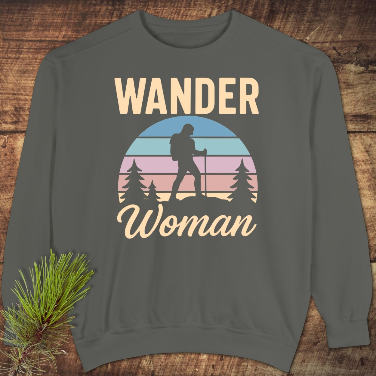 Wander Woman Retro Sweatshirt featuring a stylized sunset graphic with a hiker silhouette and WANDER Woman text, laid flat on a wooden surface with a pine sprig beside it.