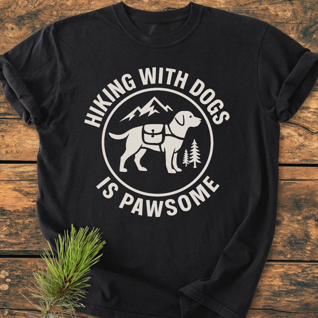 Hiking With Dogs Is Pawsome - Vintage Wash Tee featuring a dog with a hiking pack in a circular graphic, surrounded by pine trees and mountains, laid on a wooden surface with a pine branch.