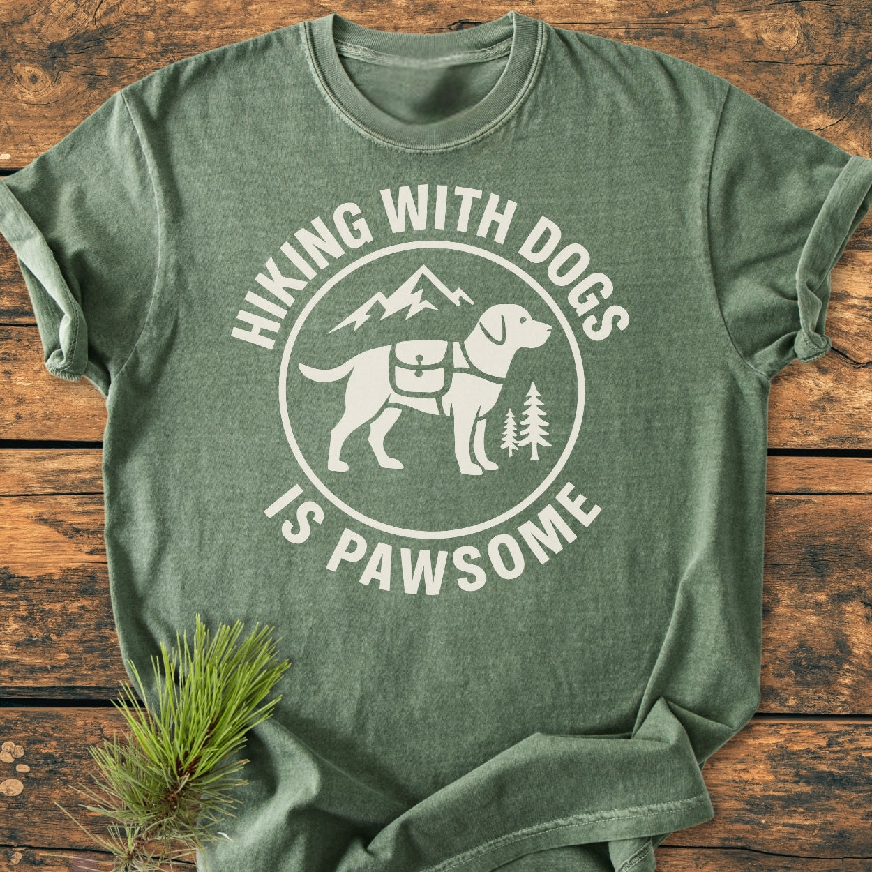 Hiking With Dogs Is Pawsome - Vintage Wash Tee features a forest-green color, showcasing a white circular badge with a dog silhouette, pine trees, and mountains, lying on a wooden surface with pine needles.