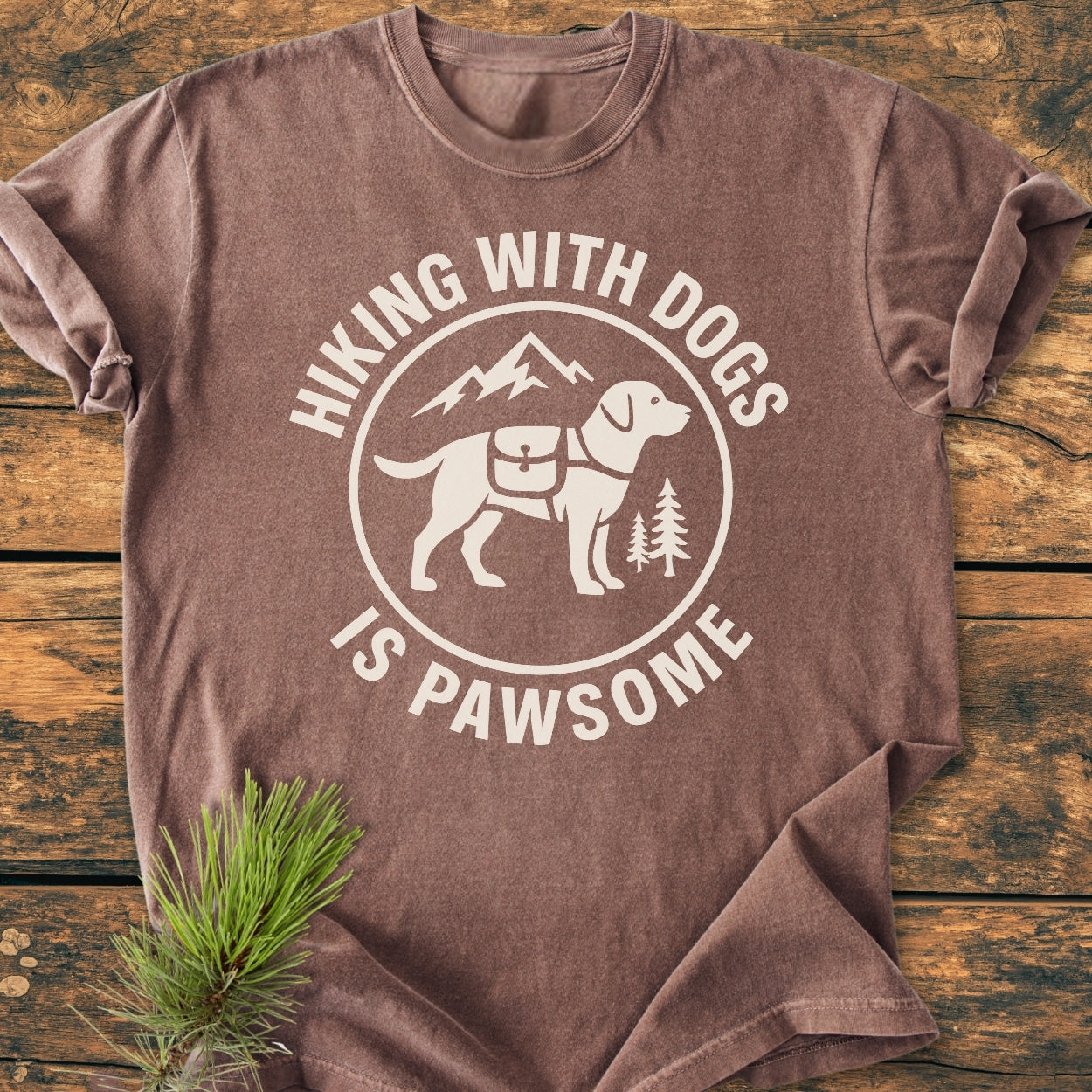 Hiking With Dogs Is Pawsome - Vintage Wash Tee featuring a hiking dog graphic with mountains and trees, laid flat on wood. Made of soft 100% ring-spun cotton for comfort and durability.