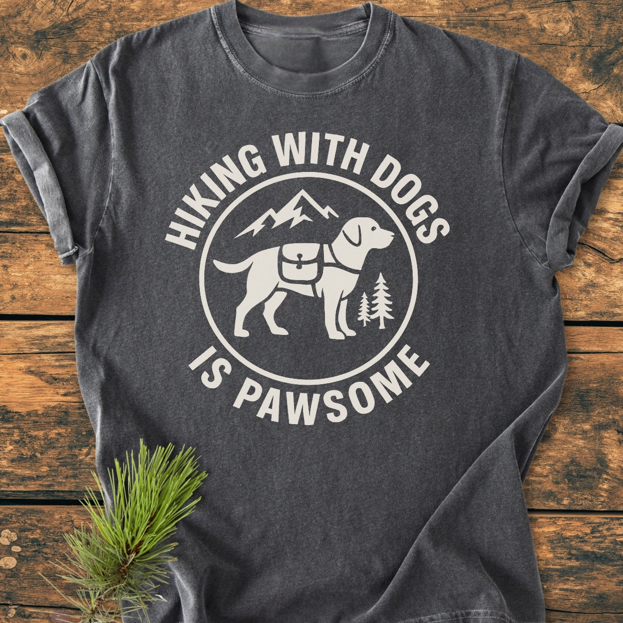 Hiking With Dogs Is Pawsome - Vintage Wash Tee features a circular logo of a dog with hiking saddlebags, pine trees, and a mountain silhouette, laid on a wooden surface with pine needles beside it.