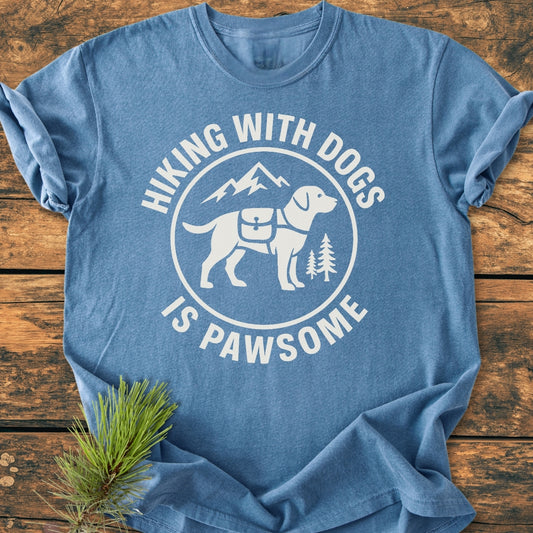 Hiking With Dogs Is Pawsome - Vintage Wash Tee featuring a front graphic of a dog with a hiking pack, surrounded by mountains and trees. Laid flat on a wooden surface, showcasing its relaxed fit.