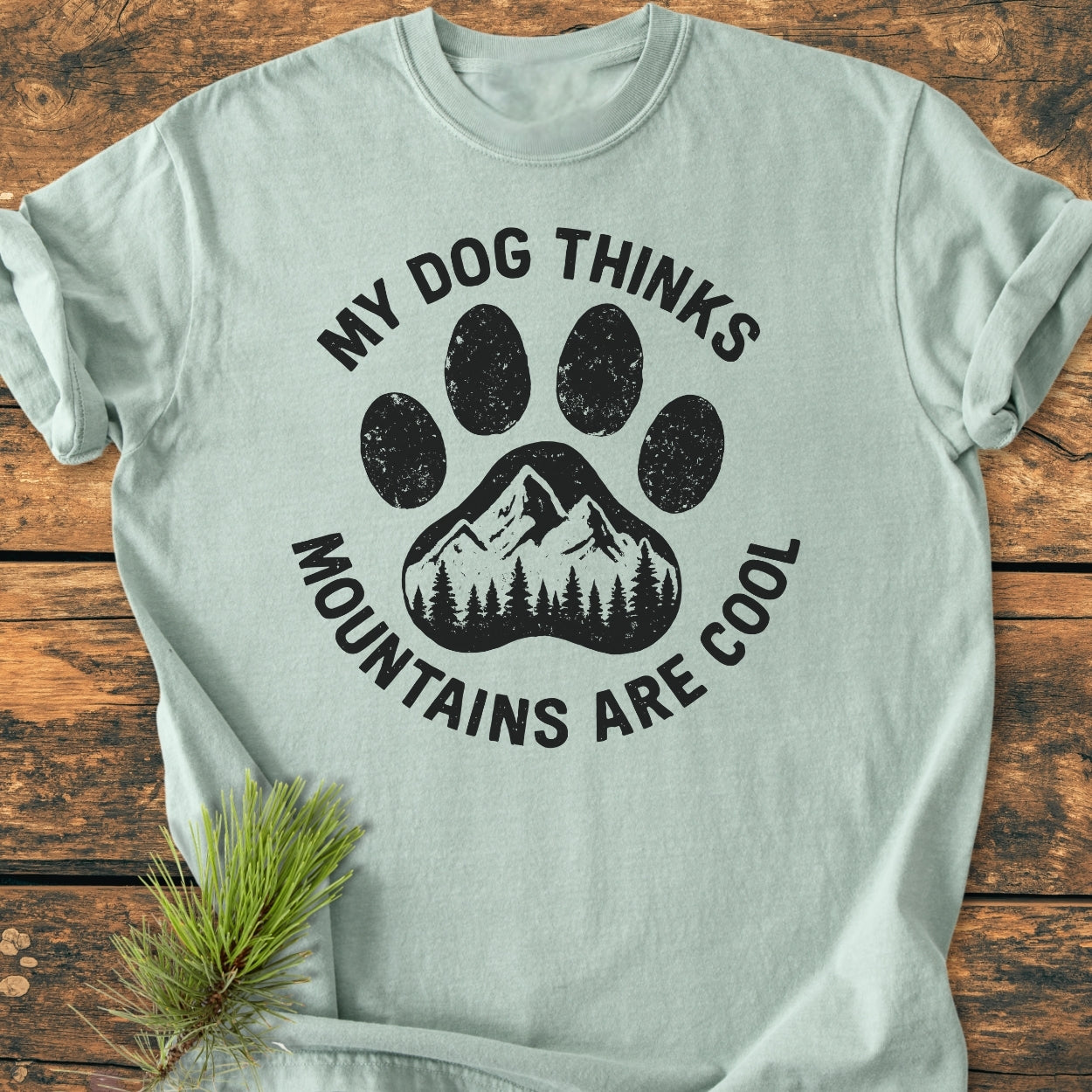 Dog Thinks Mountains Are Cool - Vintage Wash Tee featuring a paw-print with mountain and pine silhouettes, laid on wood, with a pine sprig beside. Cozy, relaxed fit, 100% ring-spun cotton.