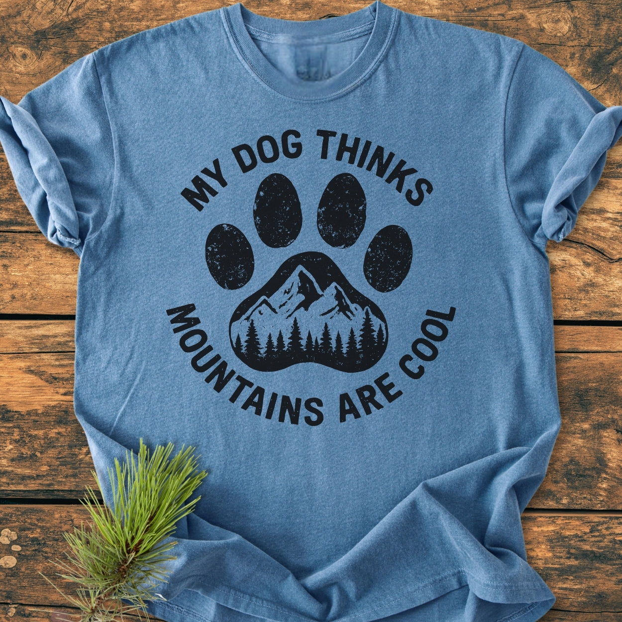 Dog Thinks Mountains Are Cool - Vintage Wash Tee features a heather-blue design with a stylized paw print and mountain silhouette, laid flat on a wooden surface, accompanied by a pine sprig.