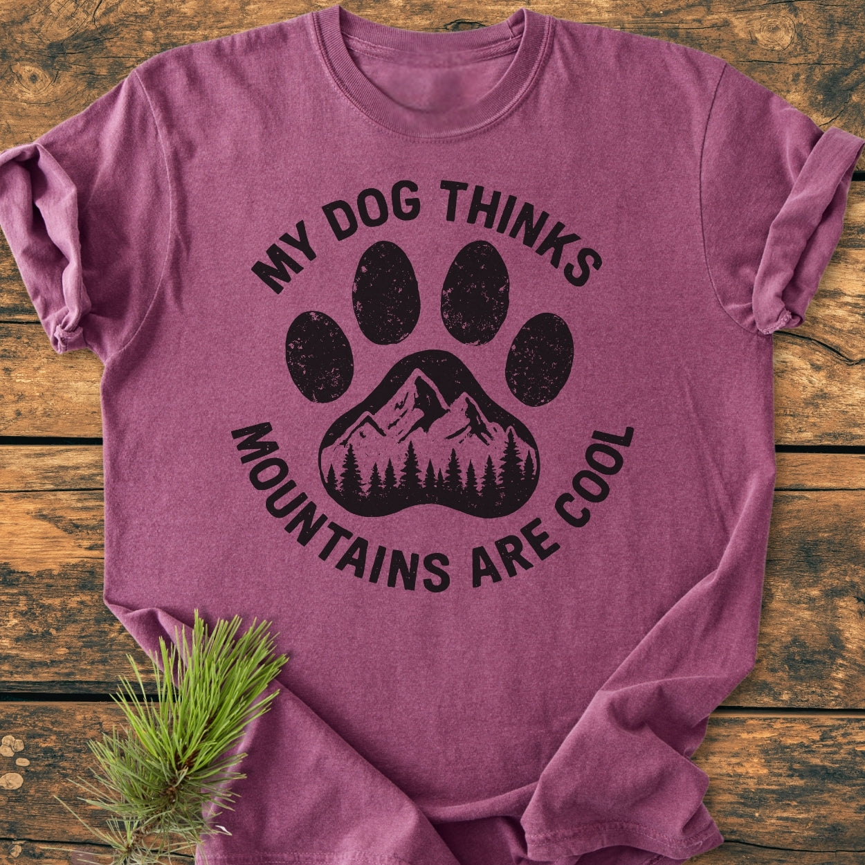 Dog Thinks Mountains Are Cool - Vintage Wash Tee features a heathered-purple color, black paw print graphic with mountains, and pine trees, displayed on wood with a pine sprig accent.