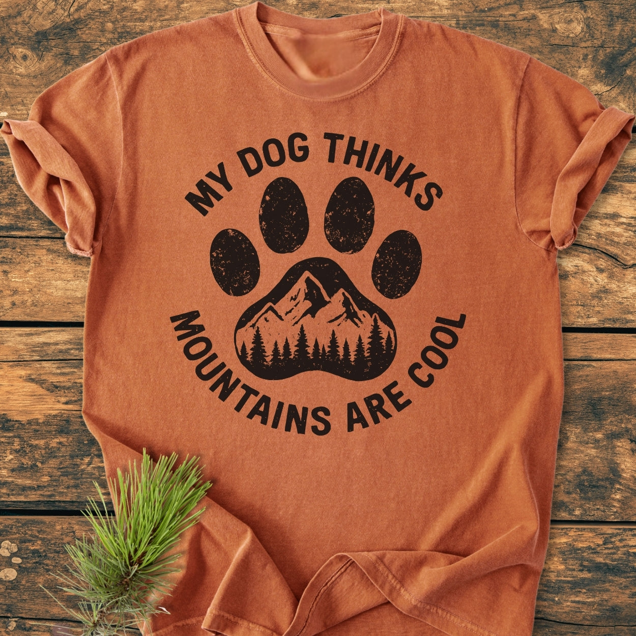 Dog Thinks Mountains Are Cool - Vintage Wash Tee features a black paw print with a mountain motif on an orange-rust backdrop, laid flat. Pine needles accent the soft, garment-dyed cotton tee.