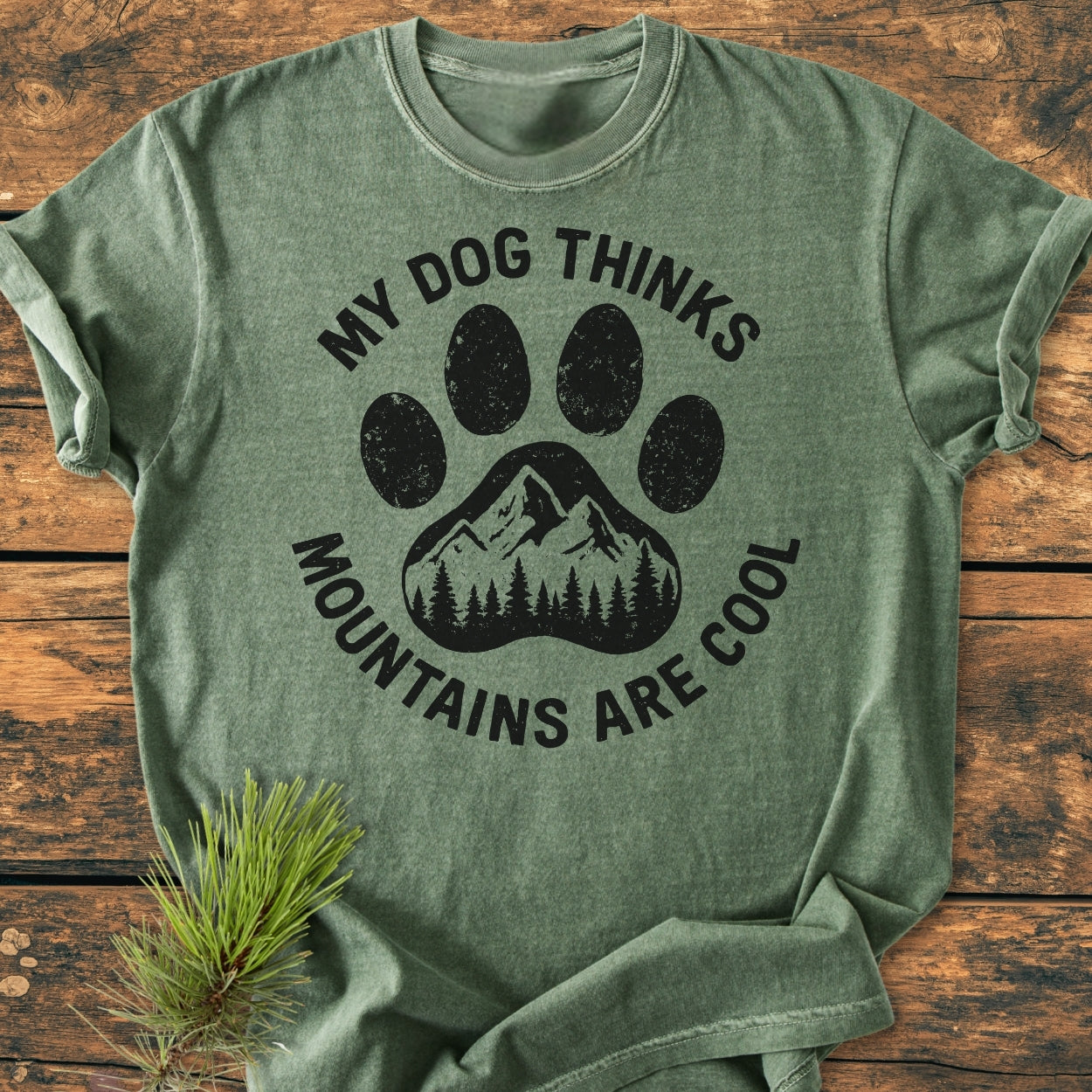 Dog Thinks Mountains Are Cool - Vintage Wash Tee features a forest-green T-shirt with a black dog-paw and mountain print, laid on wood, capturing its rustic charm and relaxed comfort.