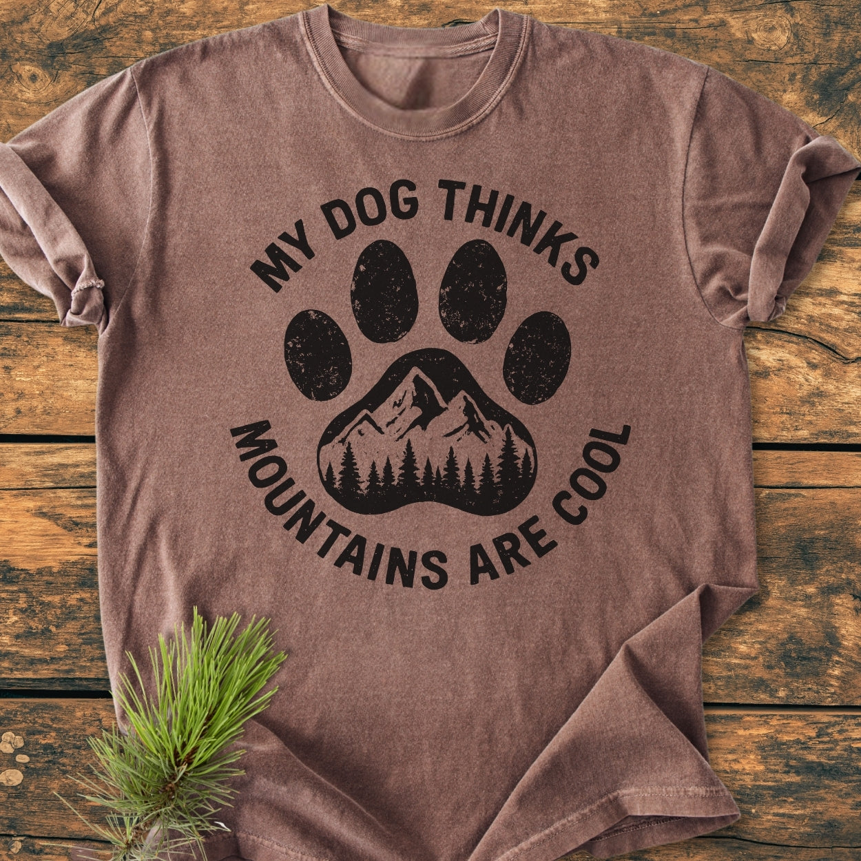 Dog Thinks Mountains Are Cool - Vintage Wash Tee featuring a large dog paw-print with a mountain range inside, laid flat on wood with a pine branch accentuating its rustic charm.
