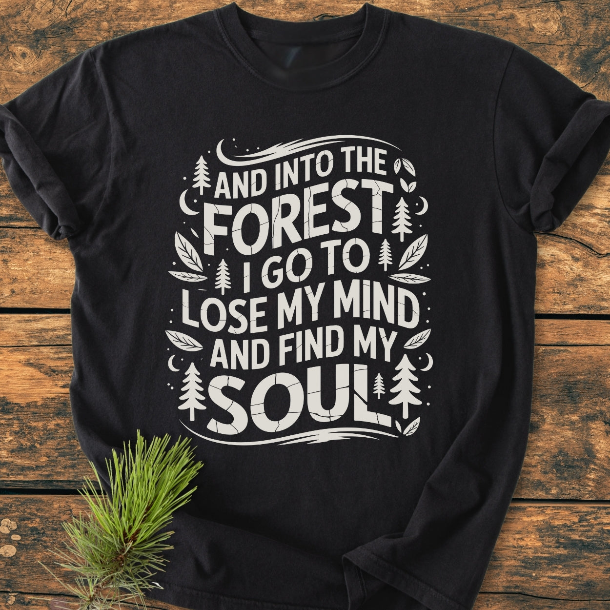 And Into The Forest I Go - Vintage Wash Tee featuring a bold white graphic with nature-themed elements, laid on wood. A soft, relaxed fit, 100% ring-spun cotton T-shirt beside a pine sprig.