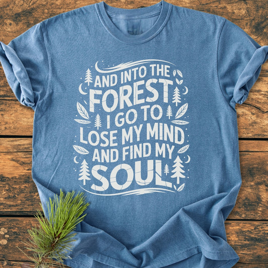 And Into The Forest I Go - Vintage Wash Tee, featuring a nature-inspired slogan with pine trees and moons, laid flat on wood. Cozy, relaxed fit made of 100% ring-spun cotton.