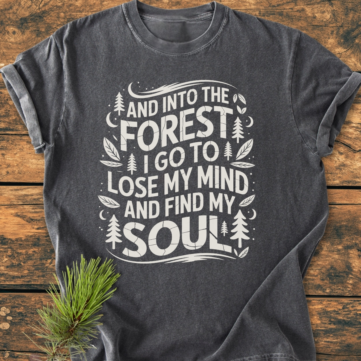 The And Into The Forest I Go - Vintage Wash Tee features a white, woodland-themed hand-lettered design on a charcoal-gray, crew-neck T-shirt, laid flat on a rustic wooden surface.