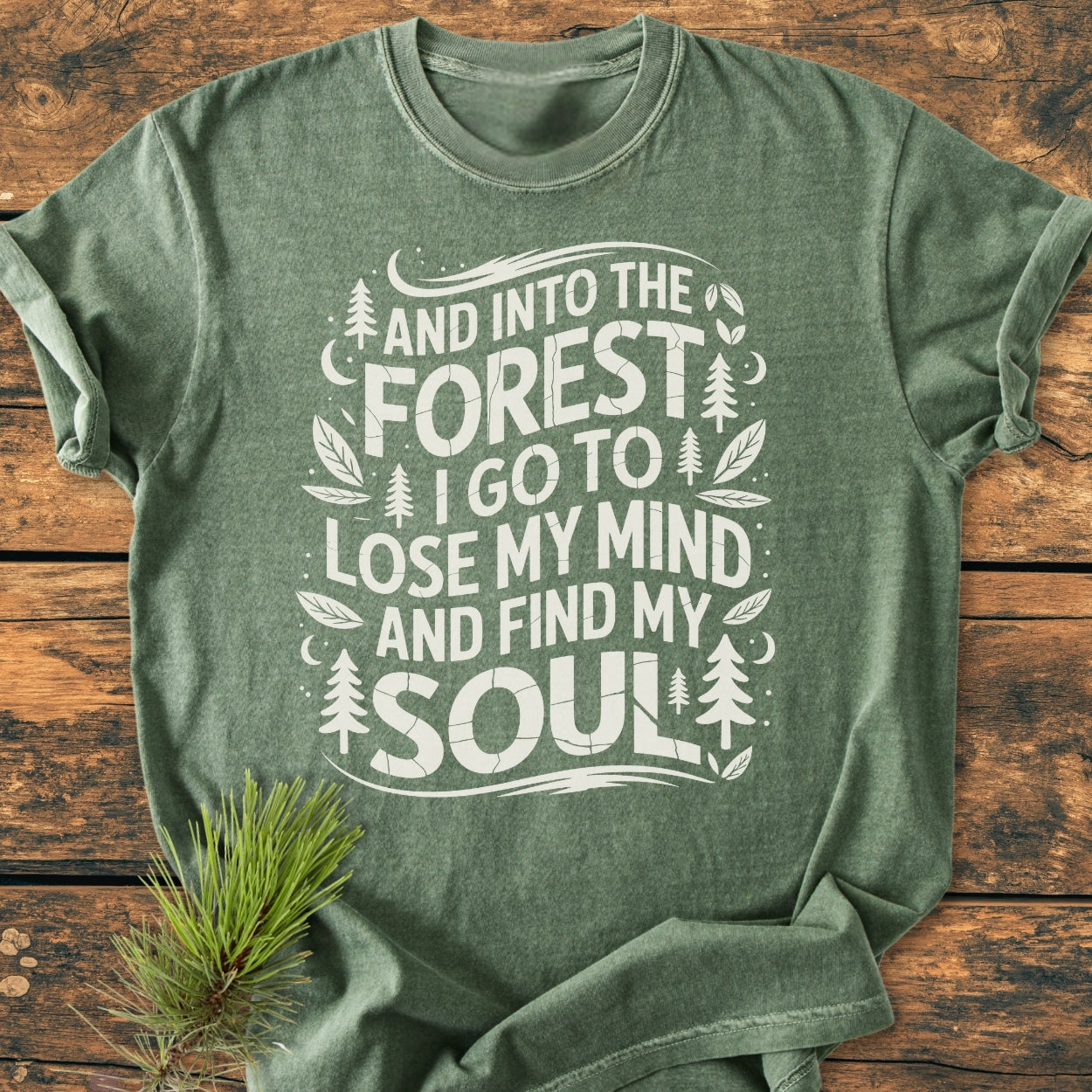 Heather-green And Into The Forest I Go T-shirt, featuring nature-inspired design with pine trees, leaves, moons, and hand-lettered quote, displayed on wood surface with small pine sprig for a rustic touch.